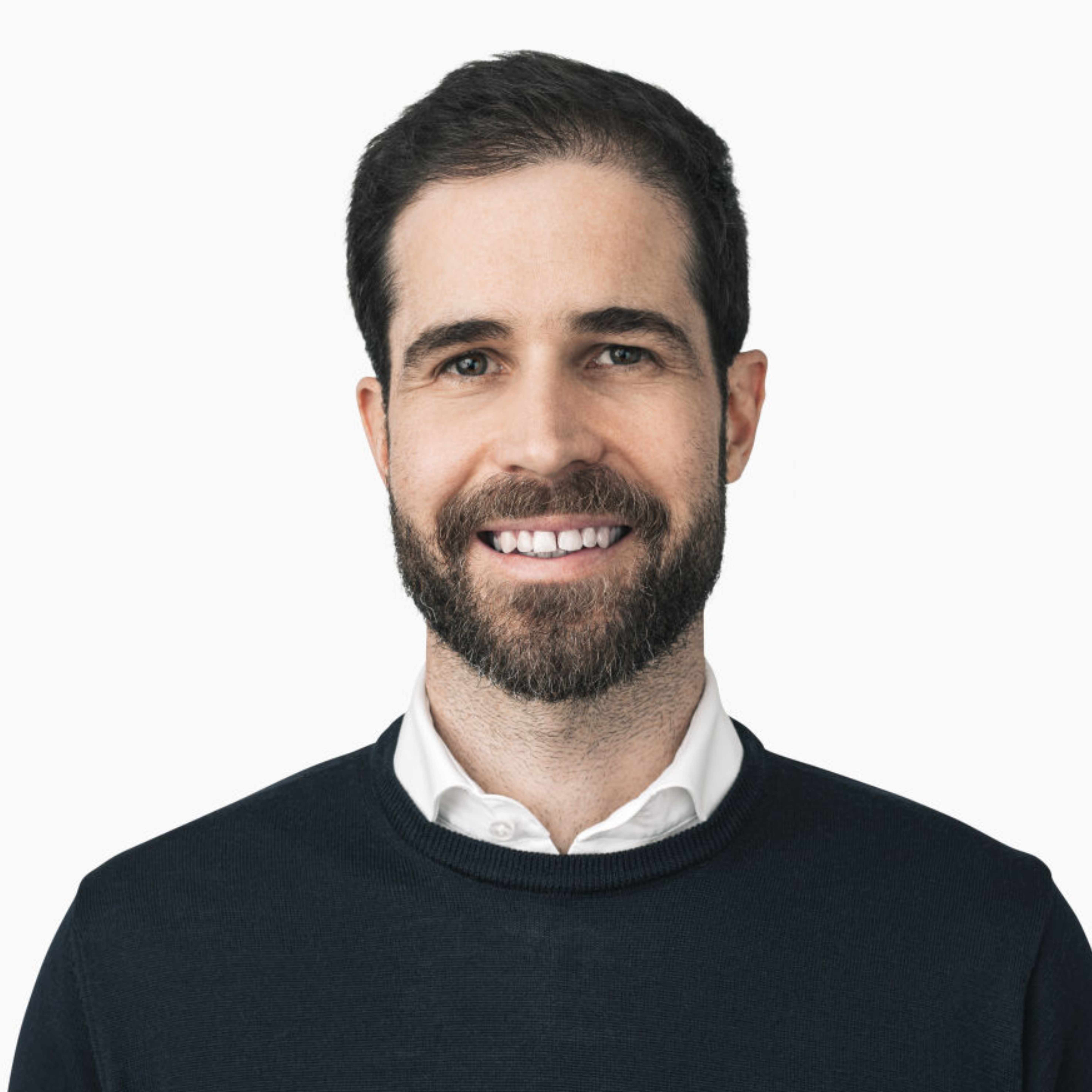 Portraitbild von Dr. Stefan Frey, einem Mann mit dunkelbraunen Haaren und einem Bart, der vor einem beigen Hintergrund fotografiert ist und ein weißes Hemd mit einem dunkelblauen Pulli darüber trägt