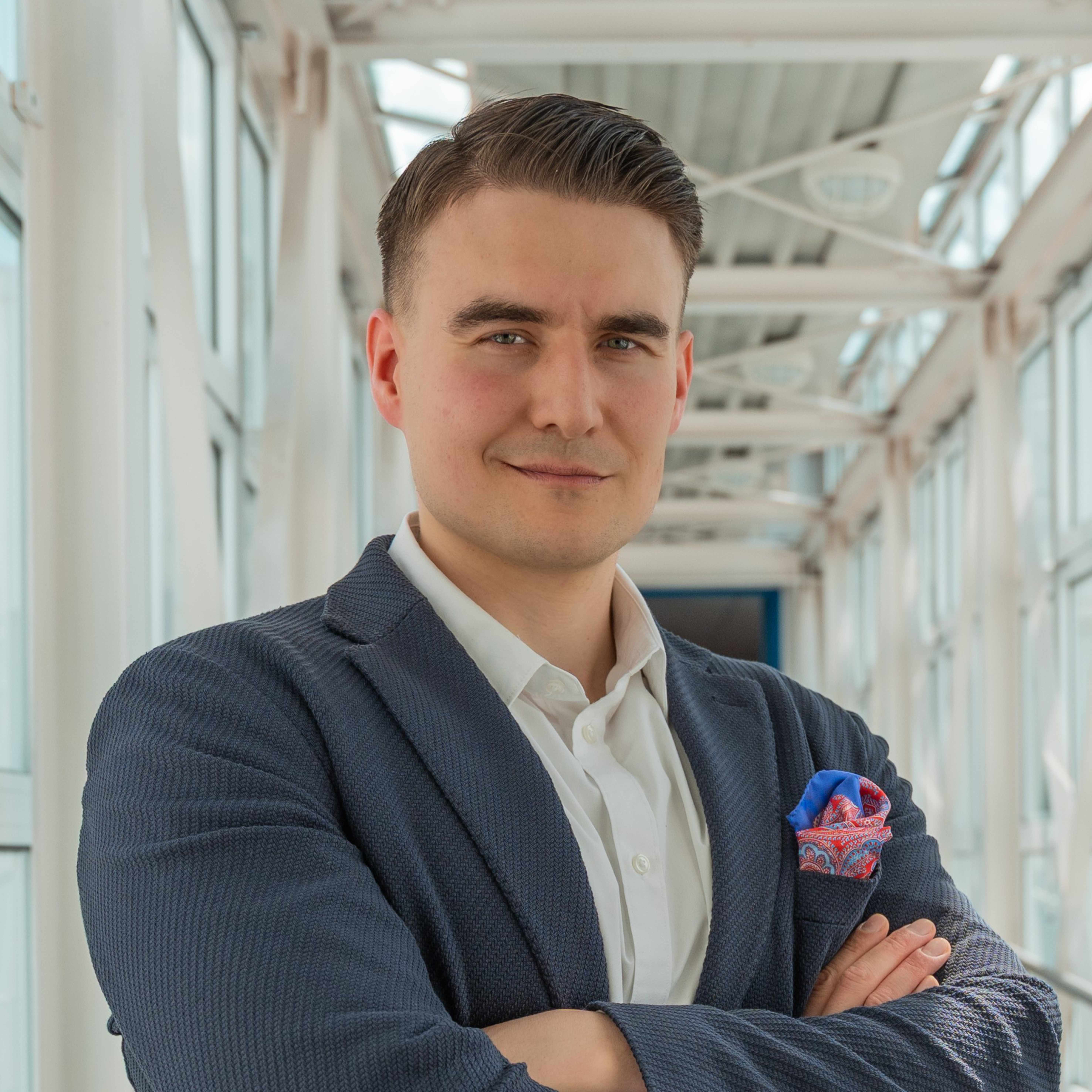 Portraitbild von Edgar Kissling, einem Mann mit dunkelbraunen Haaren, der mit verschränkten Armen in einem Gang mit weißen Stahlträgern steht. Er trägt ein weißes Hemd, ein dunkelblaues Sakko und ein rot-blau gemustertes Einstecktuch