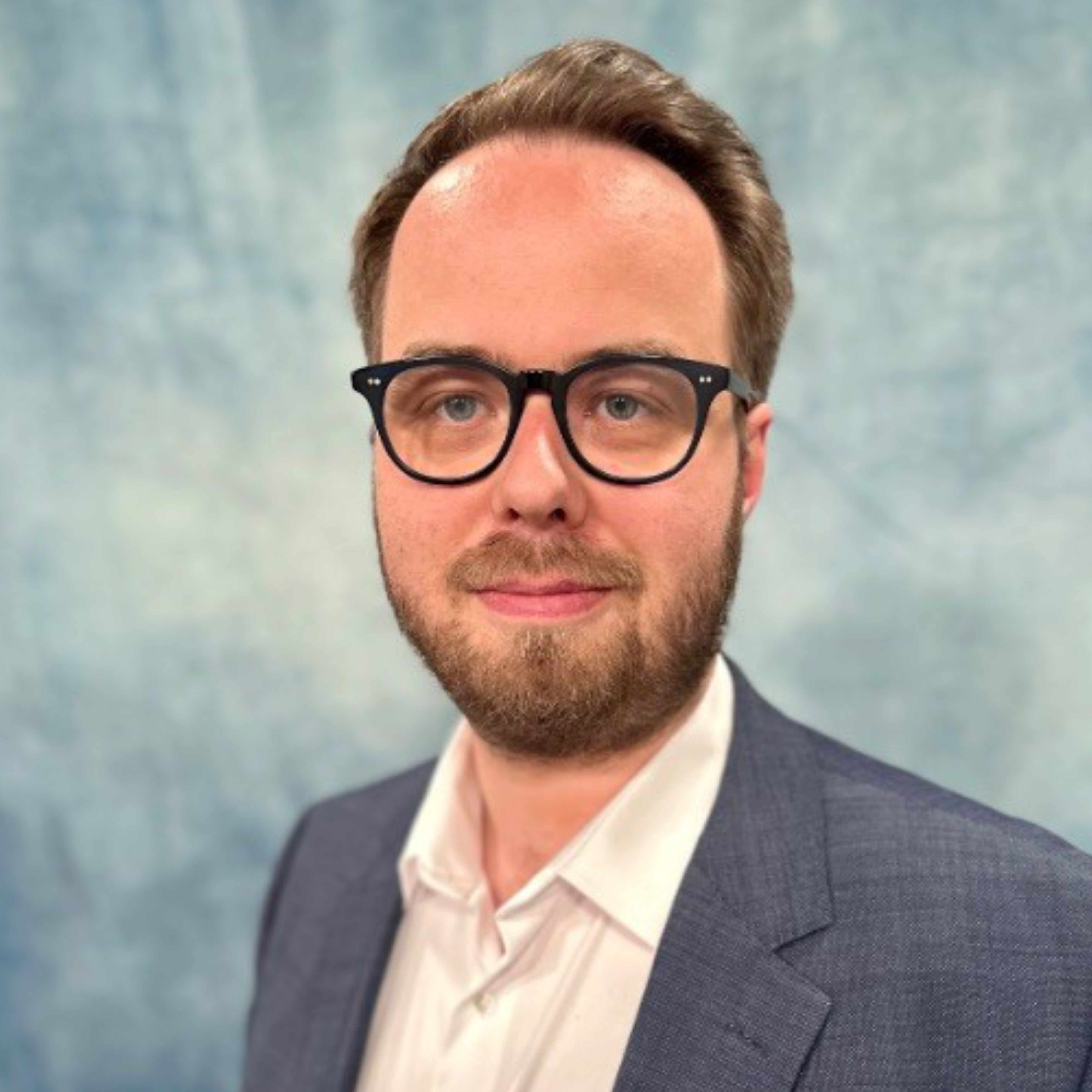 Portraitbild von Marcel Becker, einem Mann mit dunkelblonden Haare und Bart, mit einer schwarzen rundlichen Brille, der vor einem türkisfarbenen Hintergrund fotografiert ist und ein beigefarbenes Hemd mit einem blauen Sakko darüber trägt