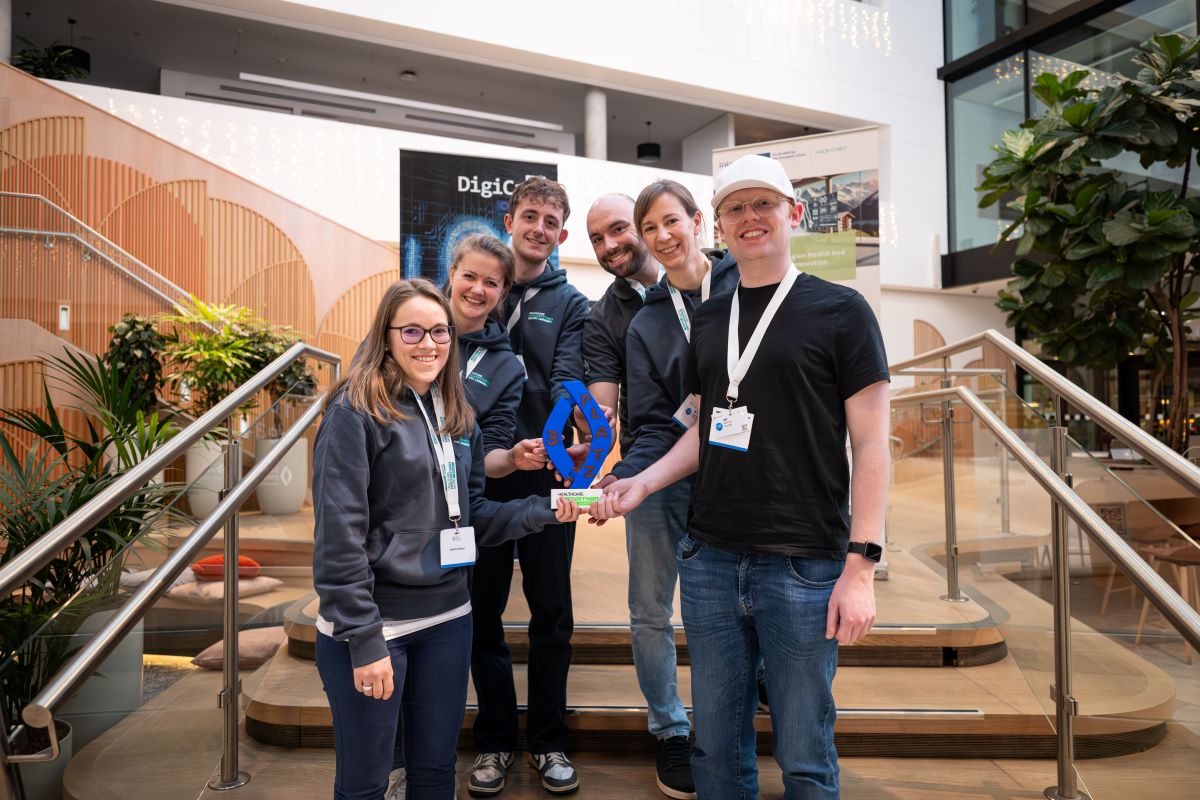 „Teilnehmende eines Hackathons stehen vor einer großen Präsentationswand und diskutieren Ideen, während im Vordergrund Arbeitsmaterialien und Laptops sichtbar sind.“