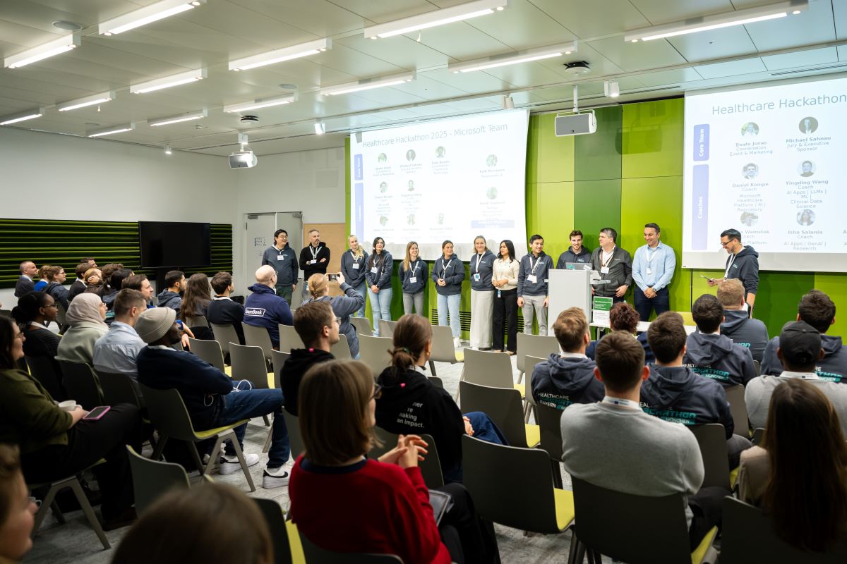 „Teilnehmende eines Hackathons sitzen in mehreren Reihen auf Stühlen und hören einer Präsentation zu, im Hintergrund ist eine große Leinwand sichtbar.“