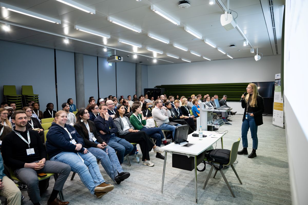 „Teilnehmende eines Hackathons sitzen in mehreren Reihen auf Stühlen und hören einer Präsentation zu, vorne ist eine große Leinwand und ein Redner sichtbar.“