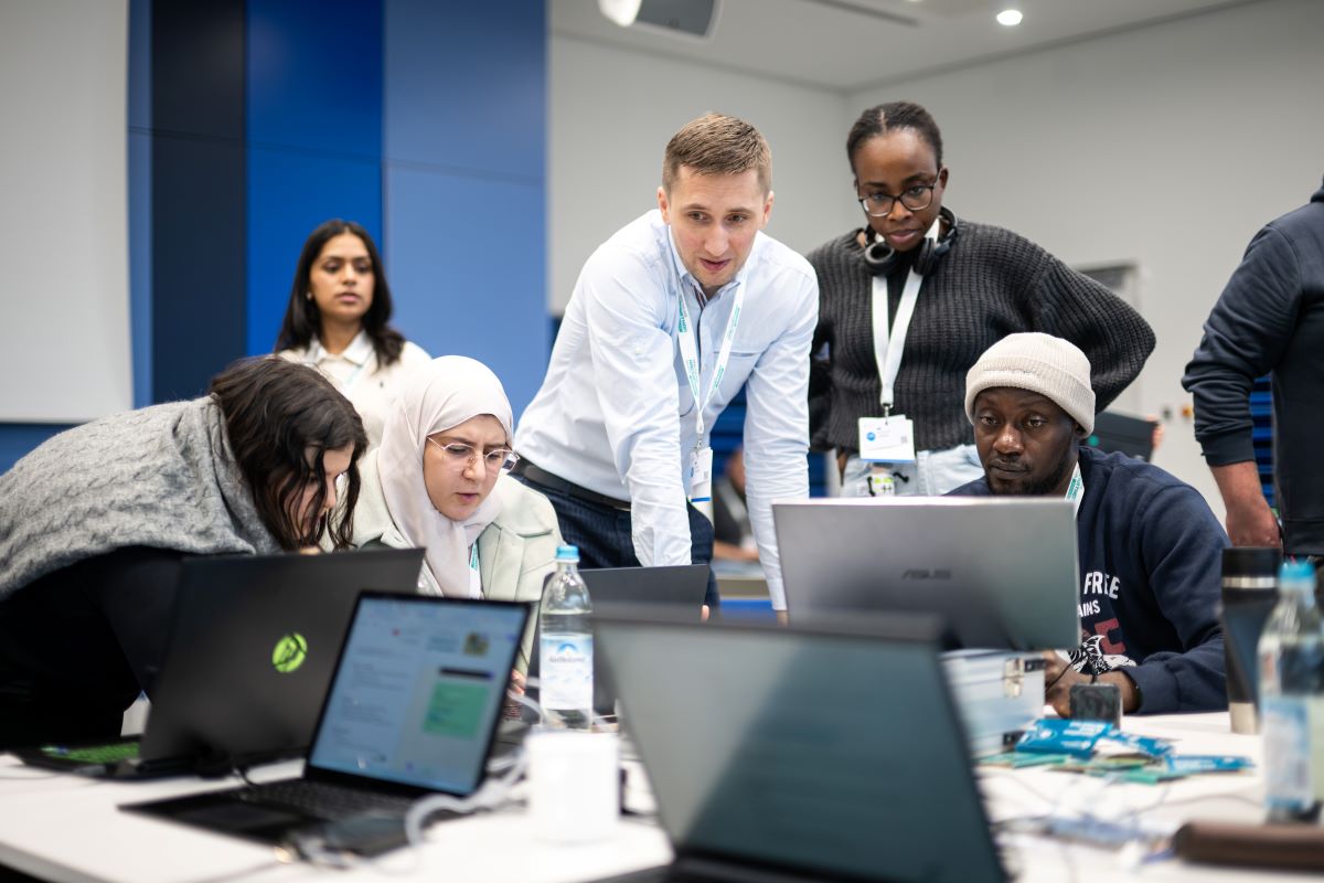 Menschen sitzen an Laptops und arbeiten, im Hintergrund stehen zwei Männer und beobachten