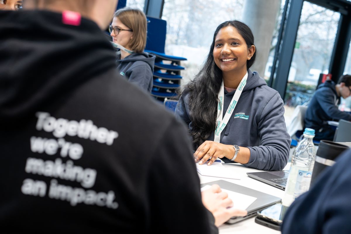 Eine Frau trägt einen Hoodie und redet mit einem Mann, der am Tisch sitzt.
