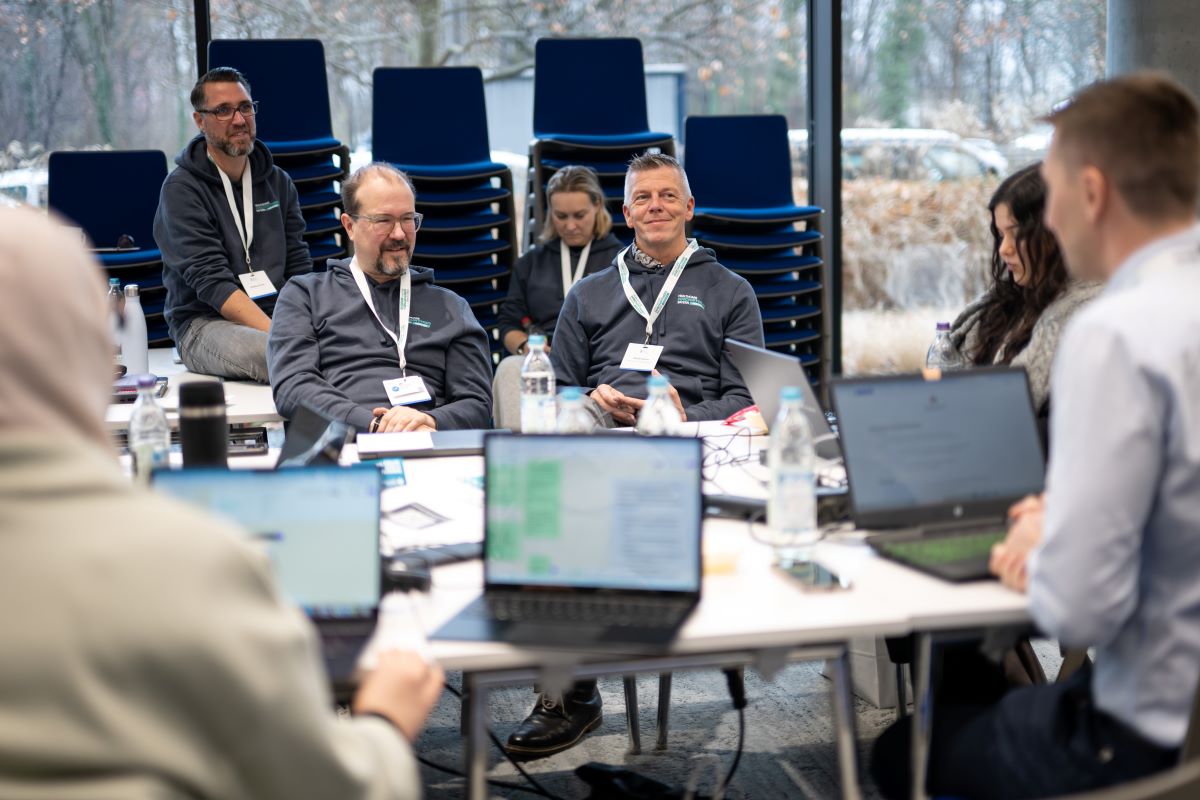 Auf dem Bild ist ein Veranstaltungsraum mit mehreren Menschen zu sehen. Einige von Ihnen sitzen vor einem Laptop.