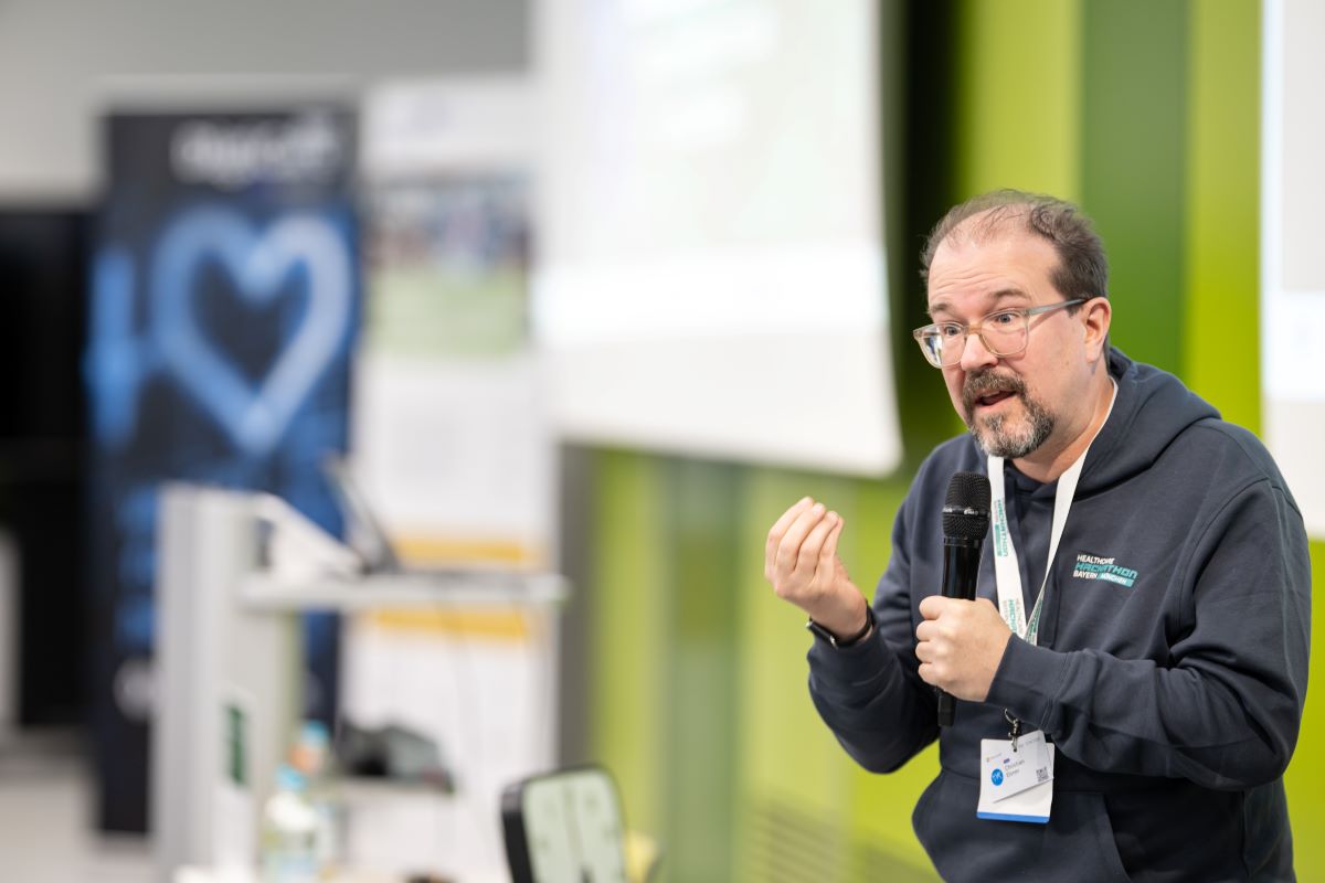 Auf dem Bild ist ein Mann zu sehen, welcher ein Mikro in der Hand hat und einen Vortrag hält. Im Hintergrund sind zwei RollUps und eine Leinwand zu sehen.