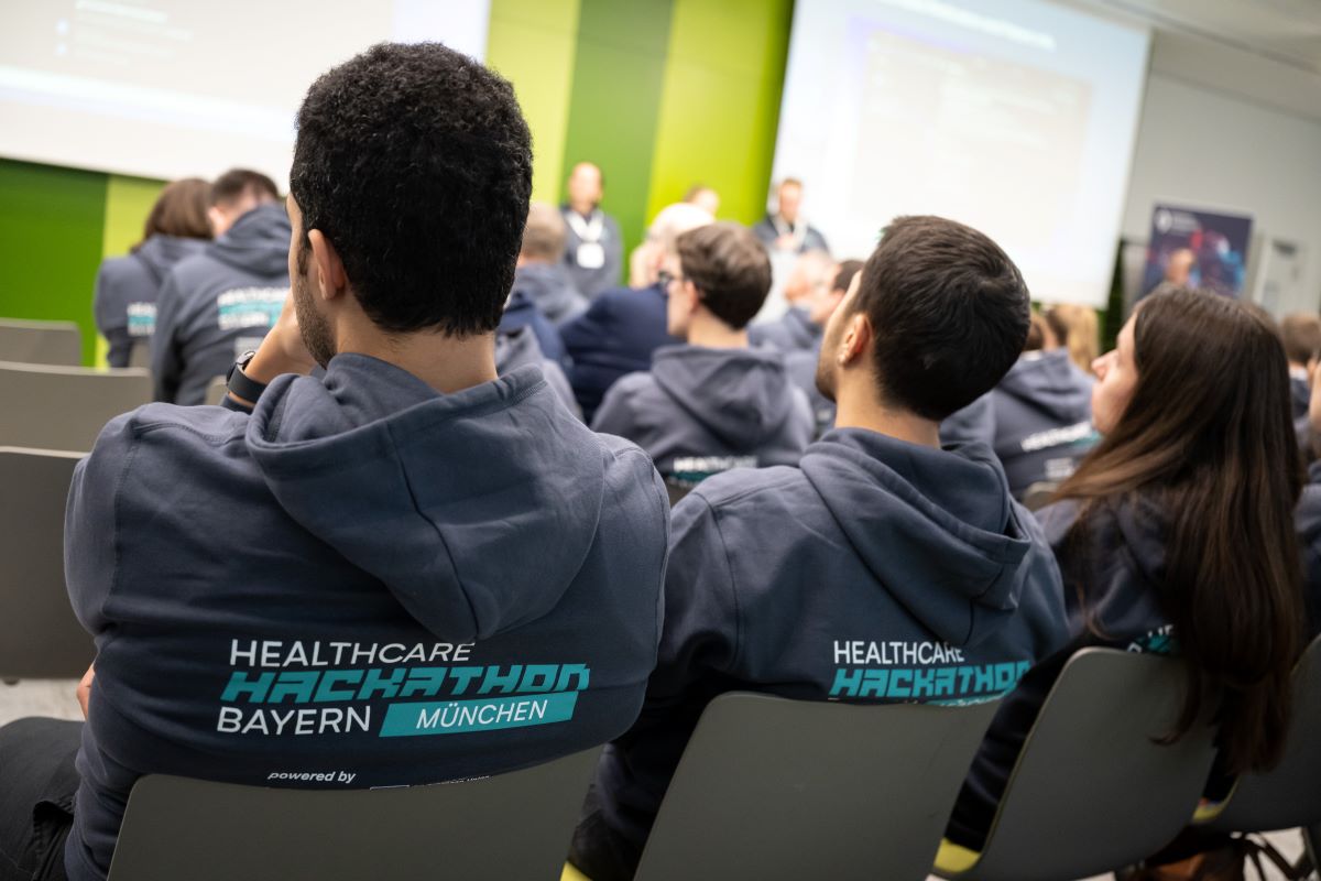 Auf dem Bild ist der Rücken von drei Menschen zu sehen, welche sich einen Vortrag anhören. Sie tragen den gleichen Pullover mit der Aufschrift Healthcare Hackathon Bayern.