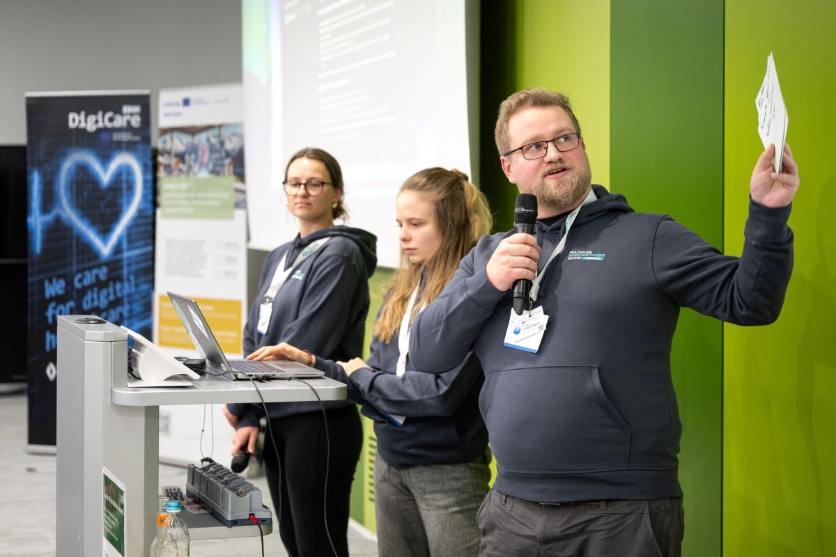 Auf dem Bild sind zwei Frauen und ein Mann zu sehen, welche alle den gleichen Hoodie tragen. Der Mann hält ein Mikrofon und Moderationskarten in der Hand. Eine der Frauen schreibt etwas am Laptop.