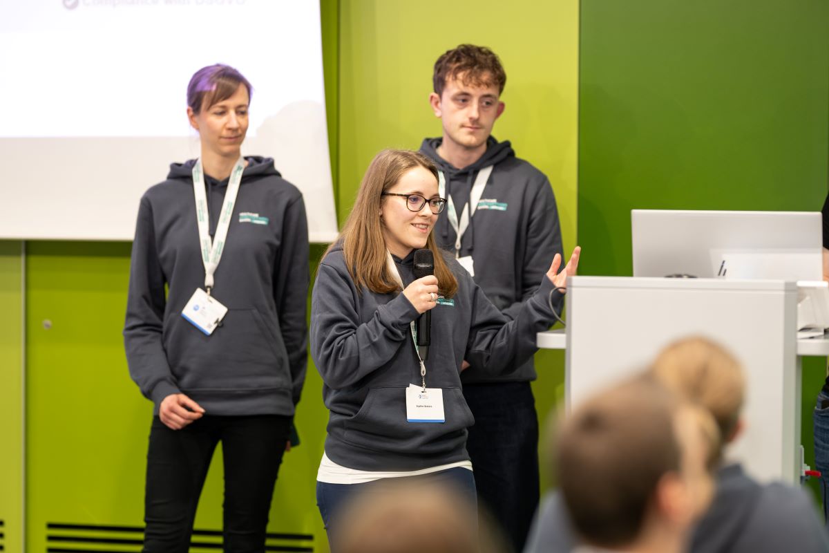 Auf dem Bild sind im Hintergrund zwei Herren in grauen Hoodies zu sehen. Im Vordergrund ist eine Dame mit Mikrofon zu sehen, welche einen Vortrag hält.