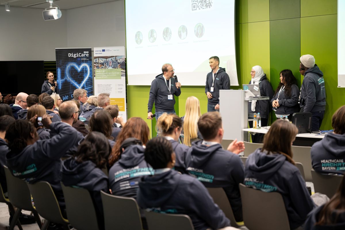 Auf dem Bild ist ein Veranstaltungsraum zu sehen, in welchem einige Personen im Publikum sitzen. Vorne stehen vor einer Leiwand ein Moderator und eine Gruppe von Menschen.