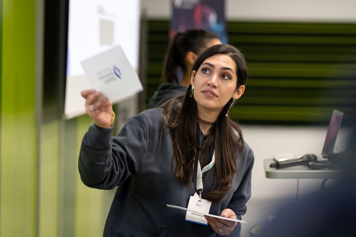 Auf dem Bild ist eine junge Frau mit schwarzen Haaren und einem dunklen Hoodie zu sehen, welche Moderationskarten in der Hand und einen Vortrag hält.