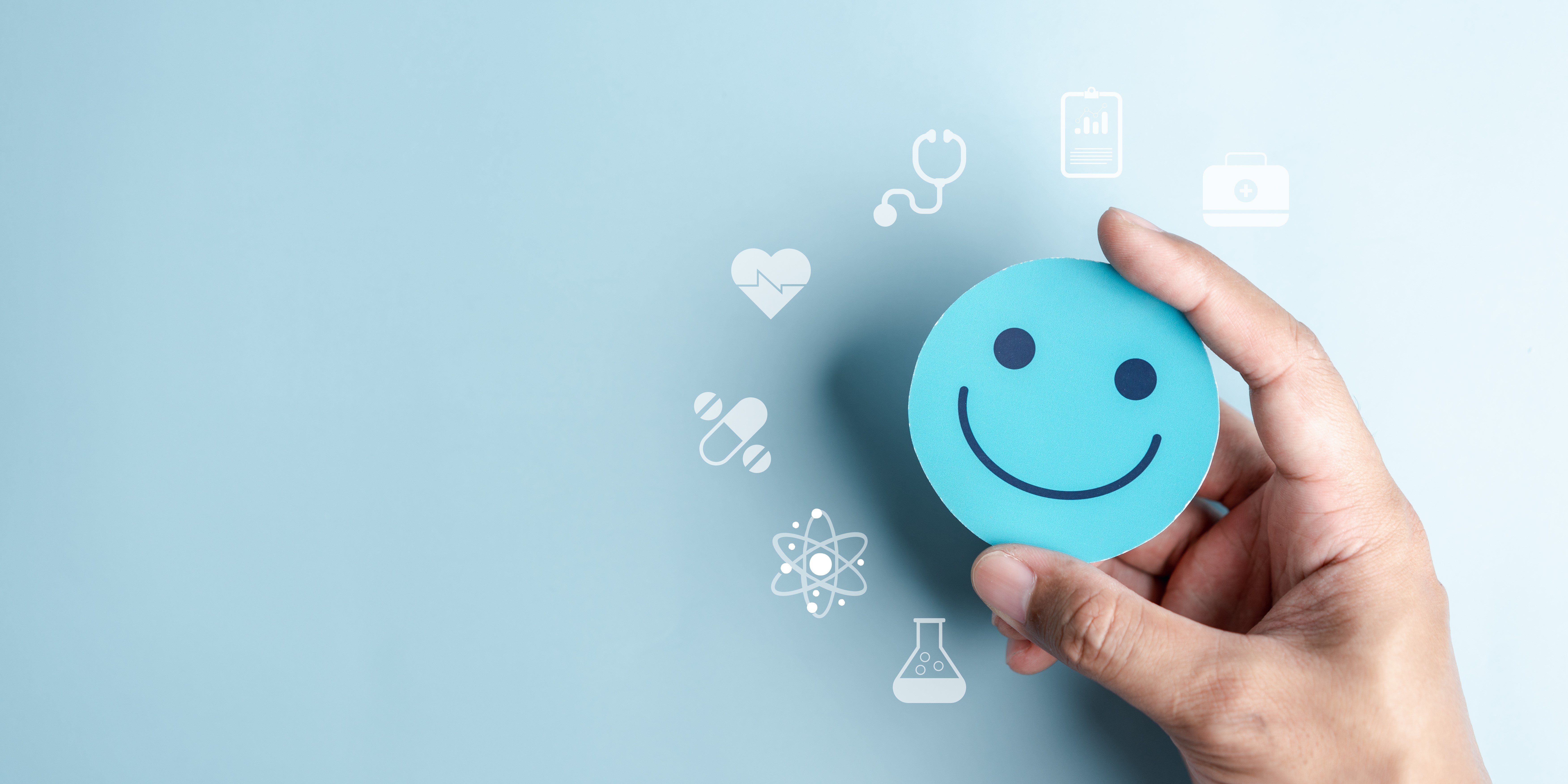Das Bild zeigt eine Hand, die ein blaues Smiley-Gesicht hält. Um das Smiley herum sind medizinische Symbole wie ein Stethoskop, ein Herz, Pillen, ein Atommodell, ein Laborkolben, eine Krankenakte und ein Erste-Hilfe-Kasten abgebildet. Der Hintergrund ist einfarbig hellblau.