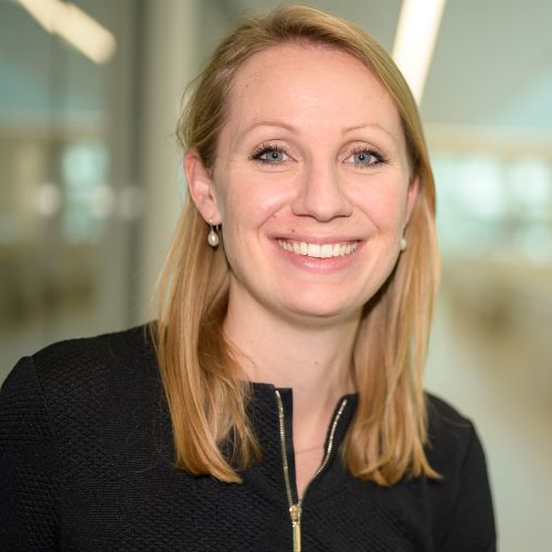 Frau mit schulterlangem, glattem Haar trägt schwarze Jacke mit Reißverschluss und Perlenohrringe vor unscharfem Hintergrund.