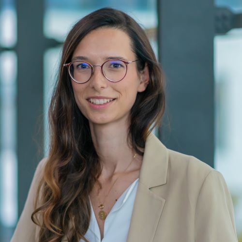 Frau mit langen braunen Haaren trägt weiße Bluse und hellbeige Blazer vor Glasfenstern