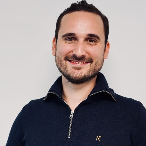 Mann mit kurzem dunklem Haar trägt einen dunkelblauen Pullover mit Reißverschluss und kleinem goldfarbenem Logo auf der Brust