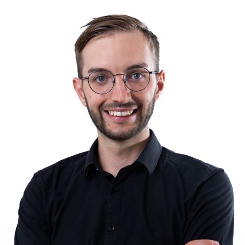 Oberkörperporträt eines Mannes mit kurzen Haaren, der ein schwarzes Hemd trägt und die Arme vor der Brust verschränkt hat