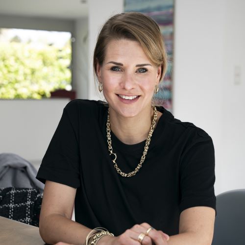 Person mit kurzem Haar, schwarzem T-Shirt und goldener Kette sitzt an einem Tisch in einem hellen Raum.