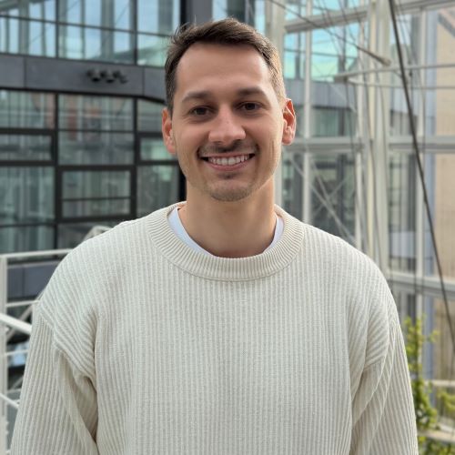 Junger Mann mit kurzem Haar trägt einen hellen, gerippten Pullover und steht vor einem modernen Glasgebäude.