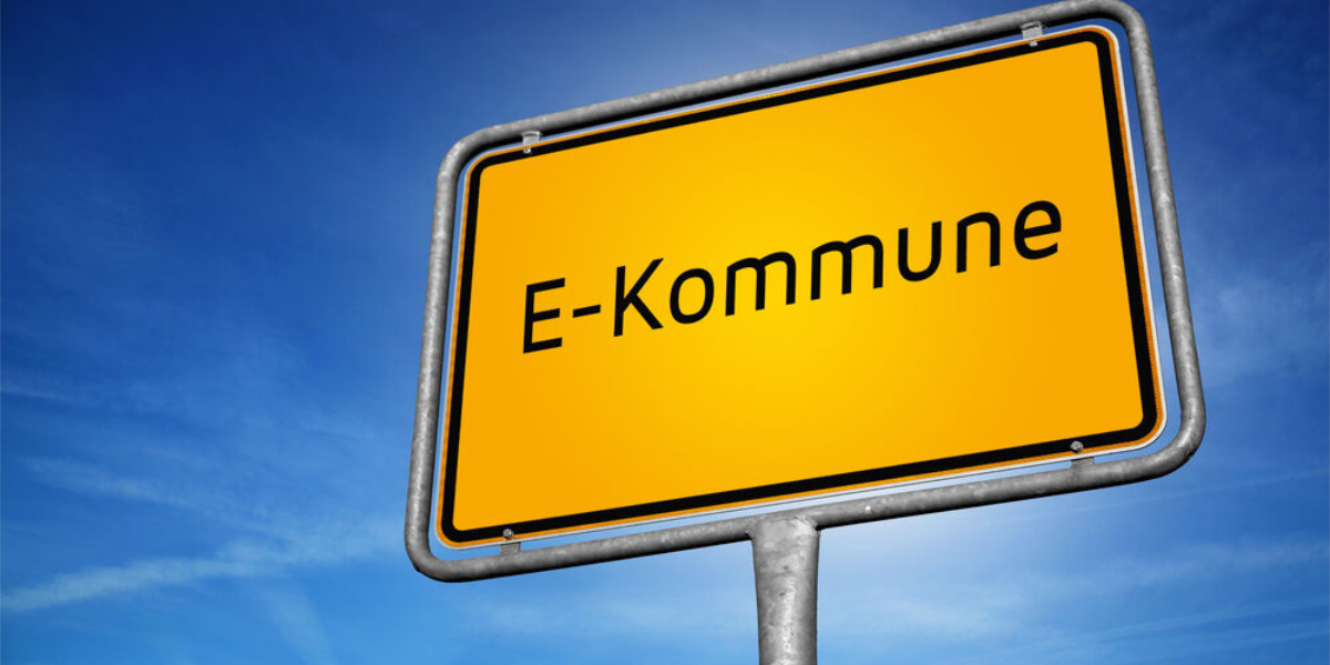 Zu sehen ist ein Ortsschild vor blauem Himmel, auf dem E-Kommune steht