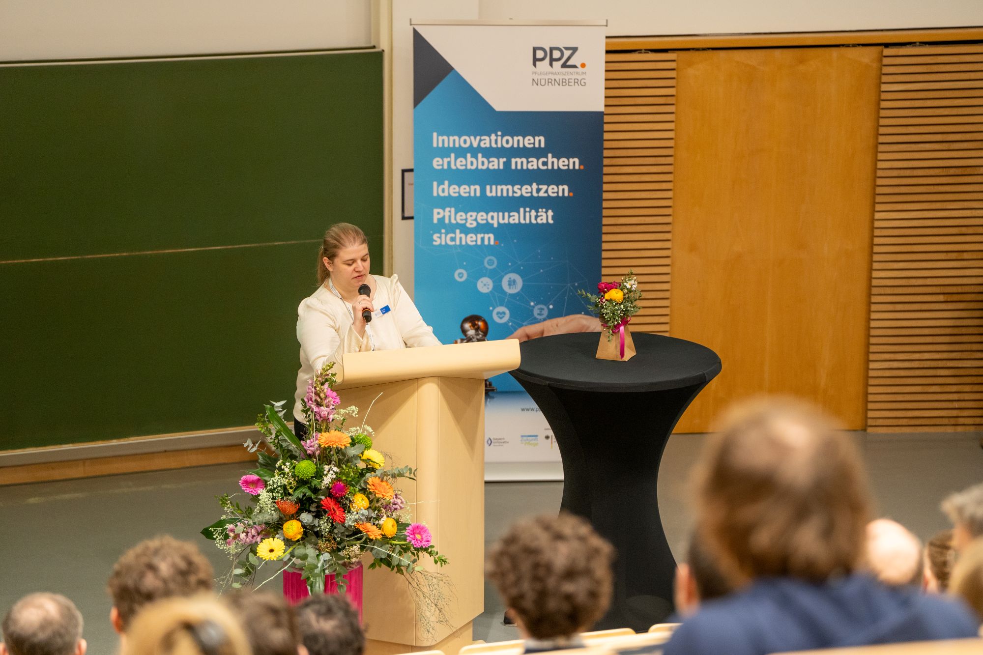 Person steht an einem Rednerpult vor Publikum in einem Hörsaal neben einem Banner mit dem Text 'PPZ NÜRNBERG Innovationen erlebbar machen. Ideen umsetzen. Pflegequalität sichern.'