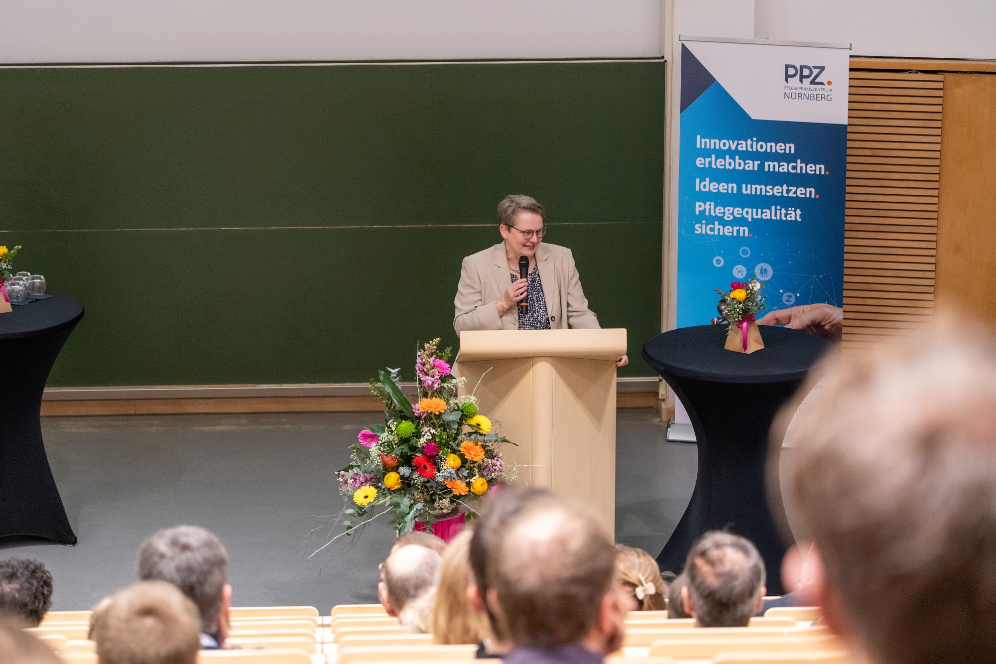 Person steht hinter einem Rednerpult vor einem grünen Tafel-Hintergrund und einem Banner mit dem Schriftzug 'PPZ NÜRNBERG Innovationen erlebbar machen Ideen umsetzen Pflegequalität sichern'