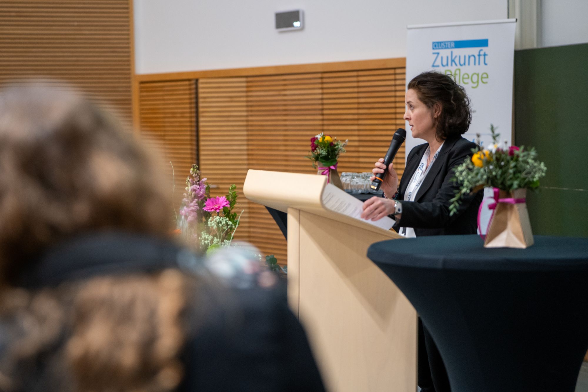 Frau steht am Rednerpult mit Mikrofon und hält Notizen, im Hintergrund Banner mit Schriftzug 'Zukunft Pflege', im Vordergrund unscharfe Person und Blumenarrangements auf Tischen