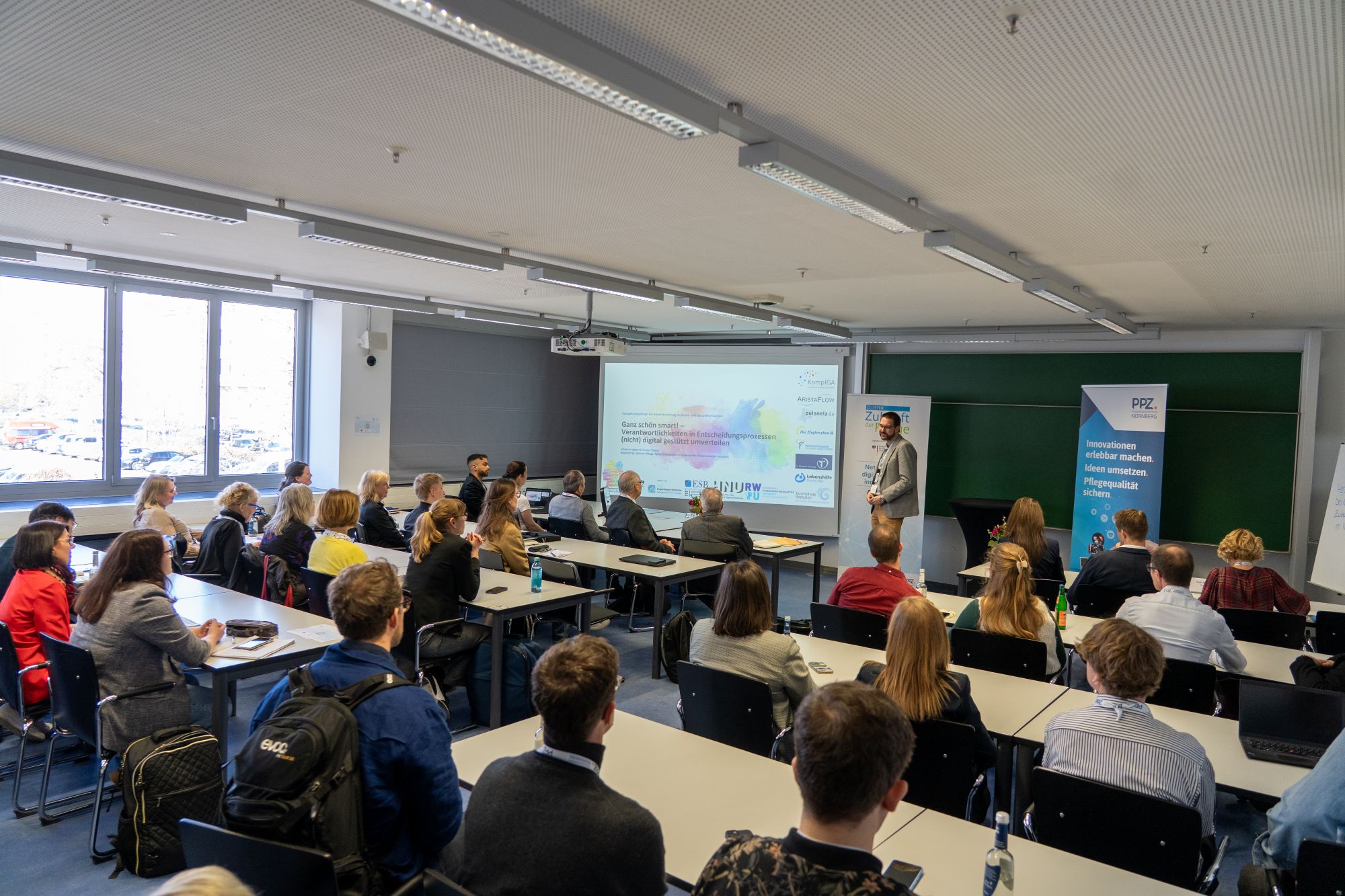 Vortragender steht vor Projektionsleinwand in einem vollbesetzten Seminarraum mit Zuhörern an Tischen