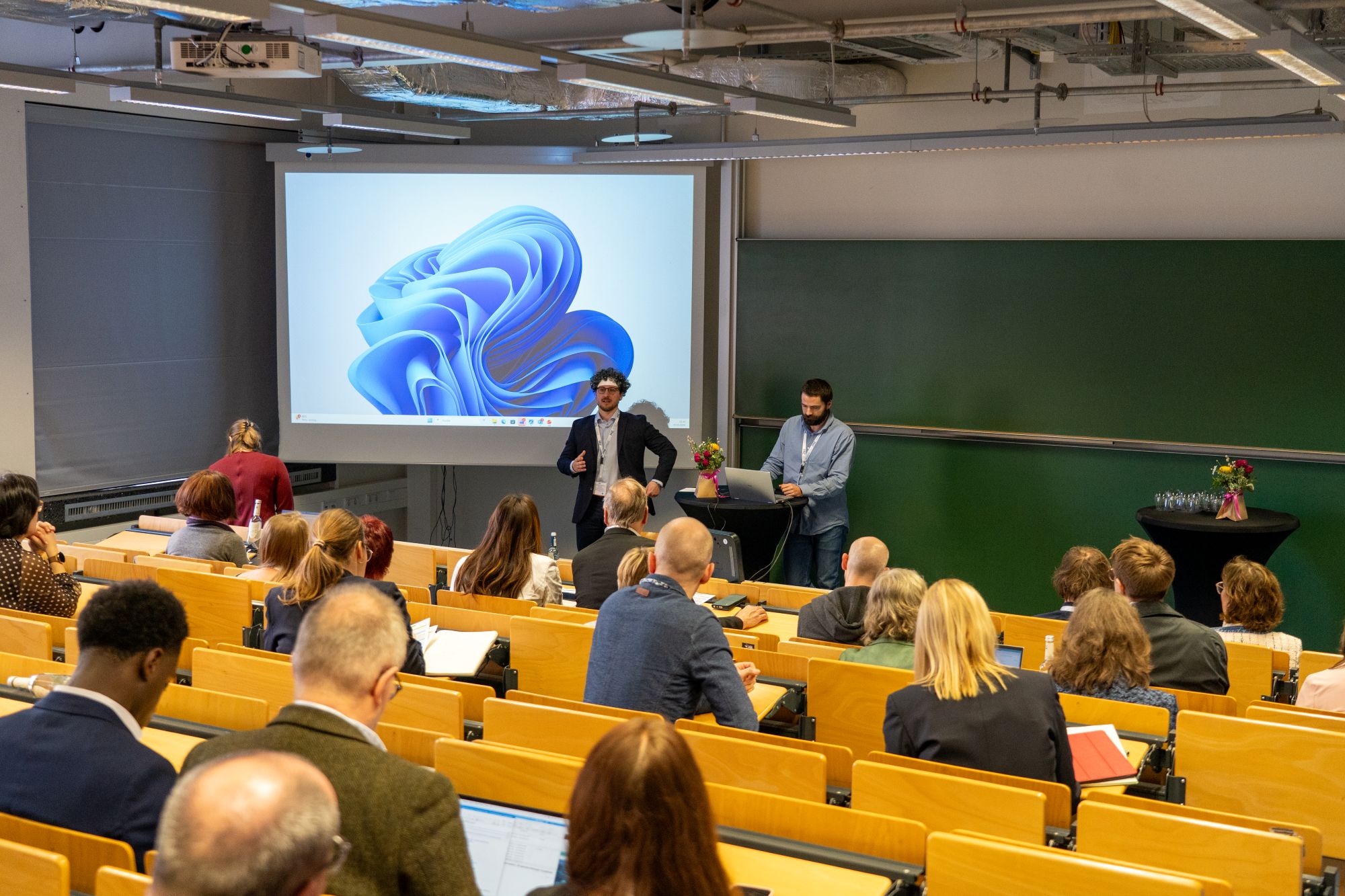 Vortragssituation in einem Hörsaal mit mehreren sitzenden Zuhörern und zwei stehenden Personen vor einer Leinwand und einer grünen Tafel