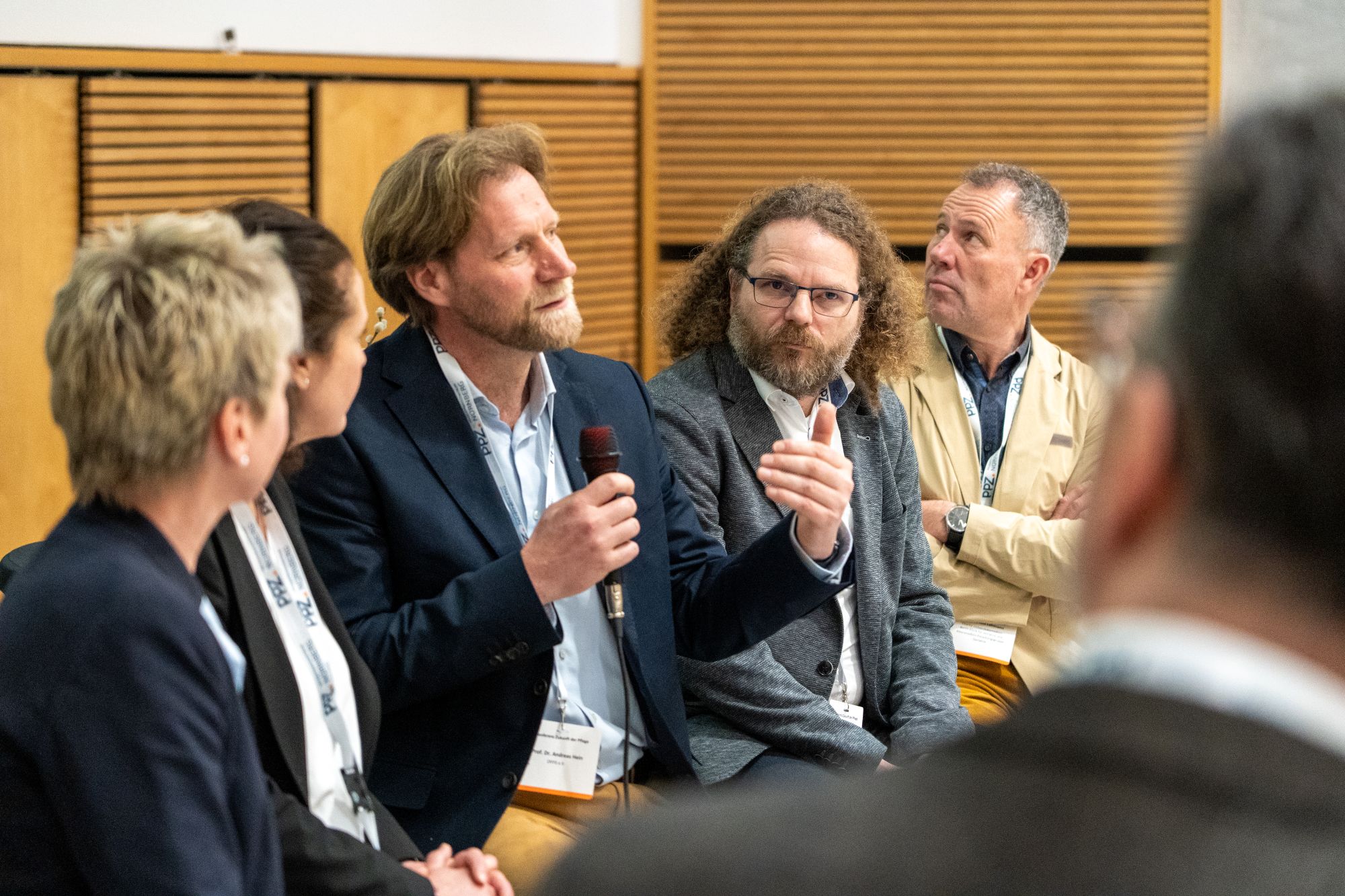Mehrere Personen sitzen in einer Diskussionsrunde, eine Person hält ein Mikrofon und spricht, im Hintergrund Holzpaneele.