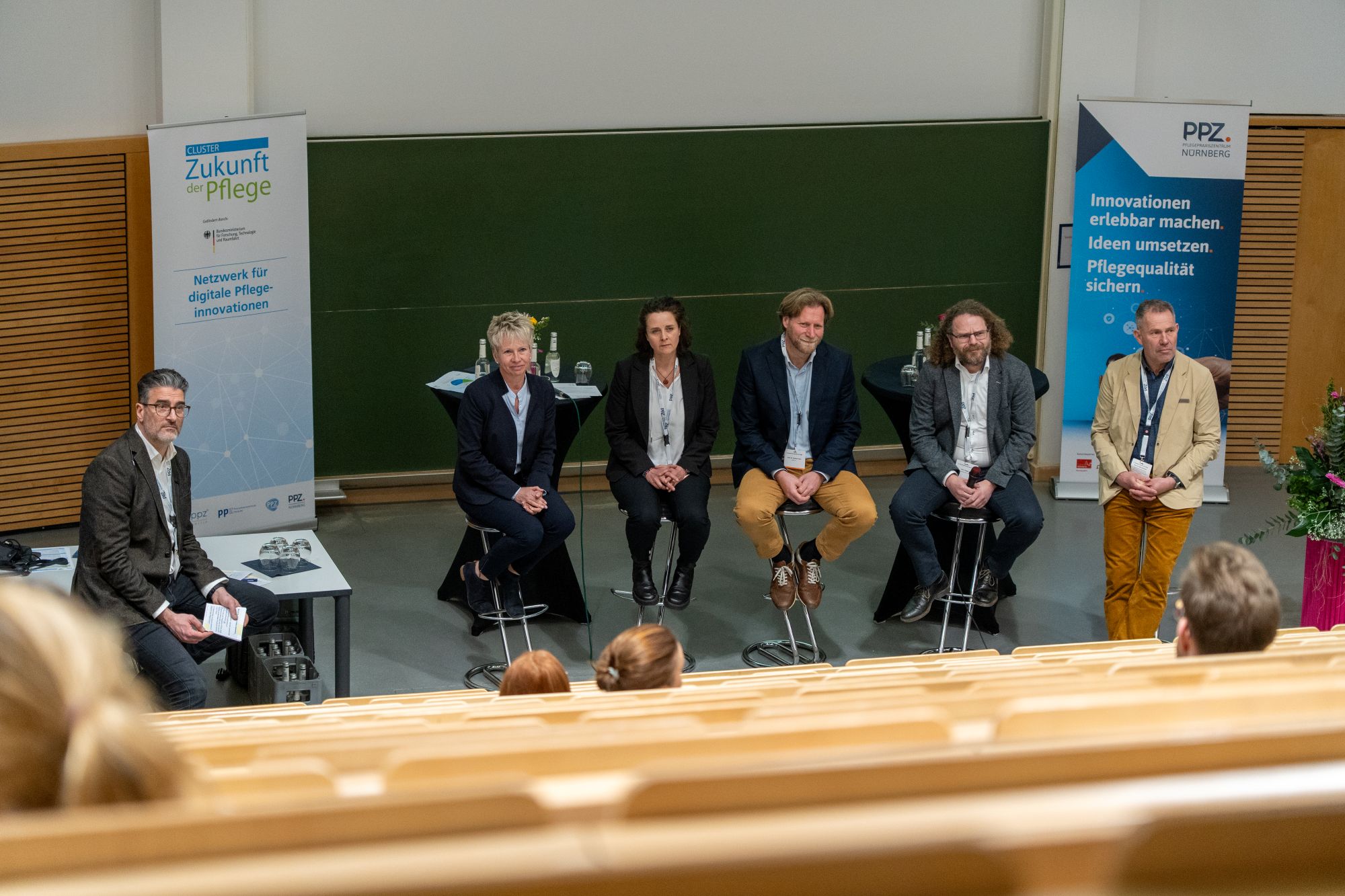 Fünf sitzende Personen auf Hockern und eine stehende Person in einem Hörsaal mit zwei Roll-ups im Hintergrund