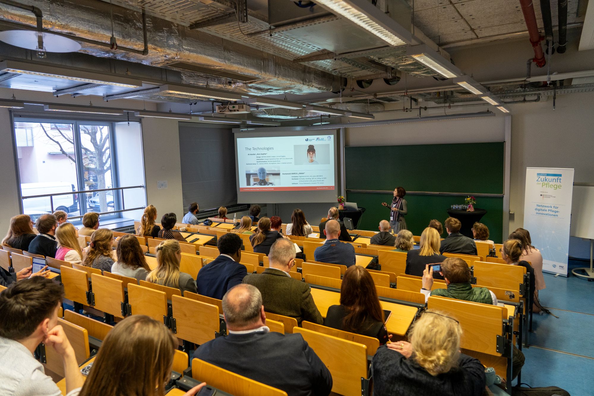 Vortragende steht vor Publikum in Hörsaal mit Projektionsfolie und grüner Tafel