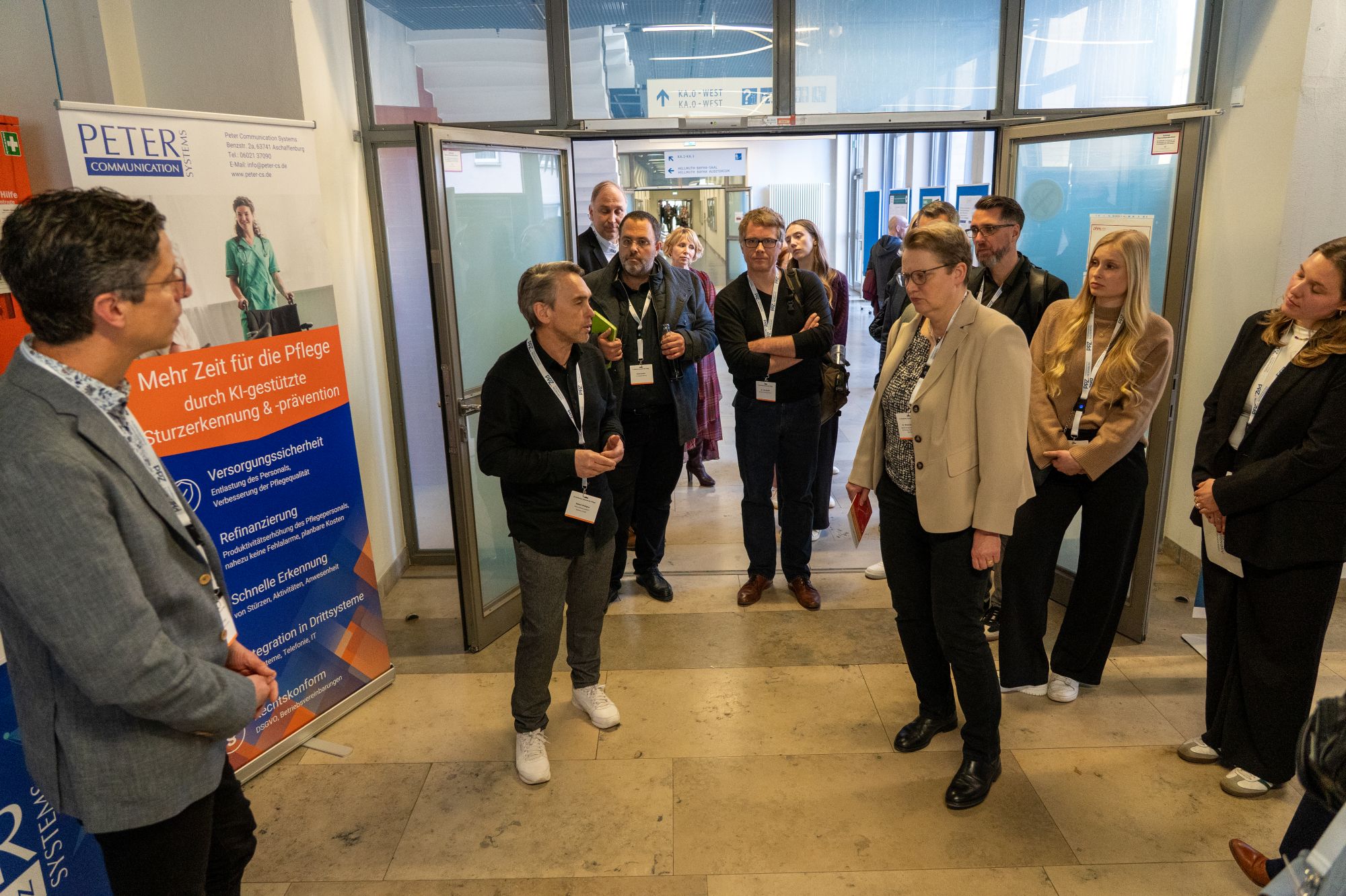 Gruppe von Menschen steht in einem Flur vor einem Informationsbanner mit Text zu Pflege und Gesundheitsversorgung
