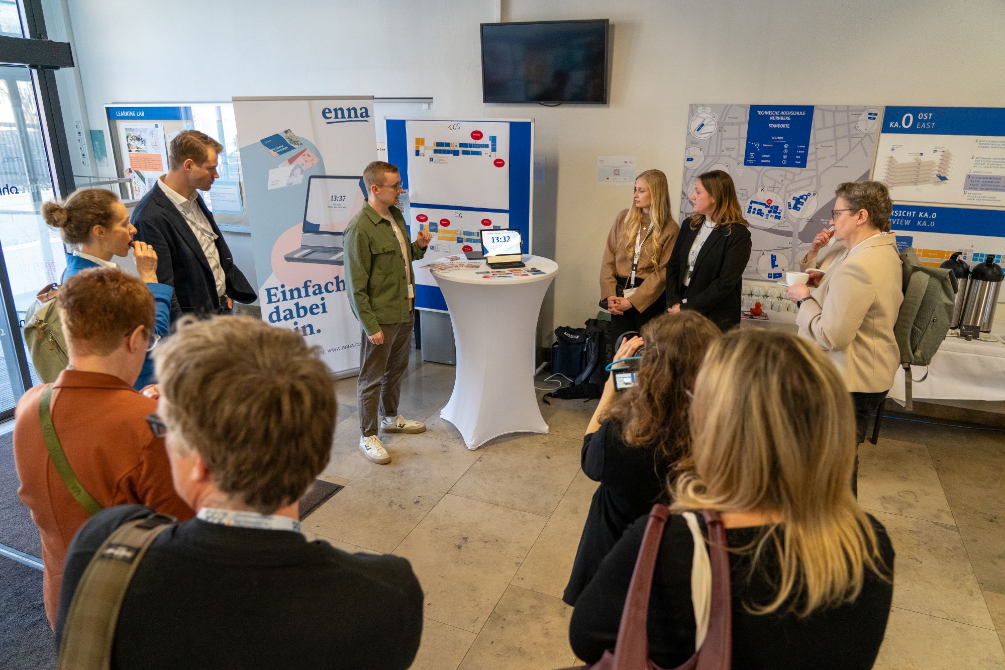 Gruppe von Menschen steht in einem Raum vor einem Stehtisch mit Informationsmaterial und Plakaten an der Wand