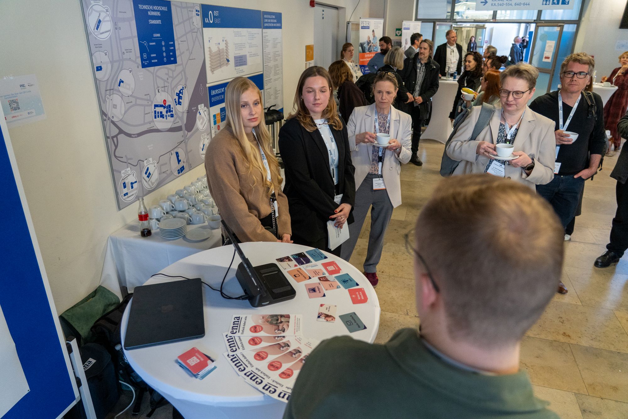 Mehrere Personen stehen an einem runden Stehtisch mit Informationsmaterial und Mikrofon in einem hellen Raum mit Plakaten an der Wand