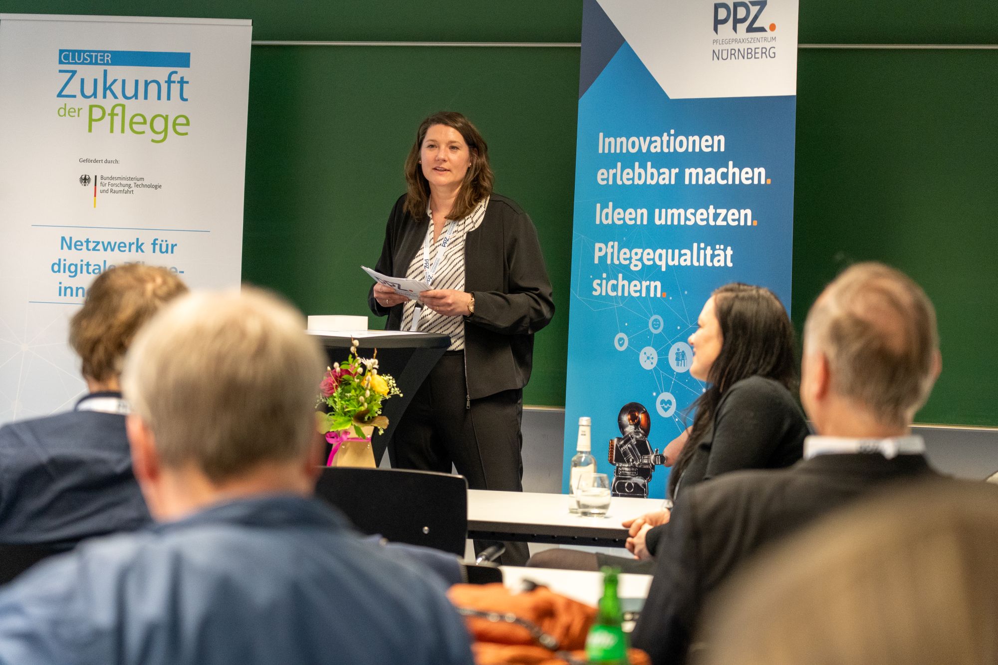 Frau steht vor Publikum und hält Papiere in einem Raum mit Bannern zu Pflege und Innovationen