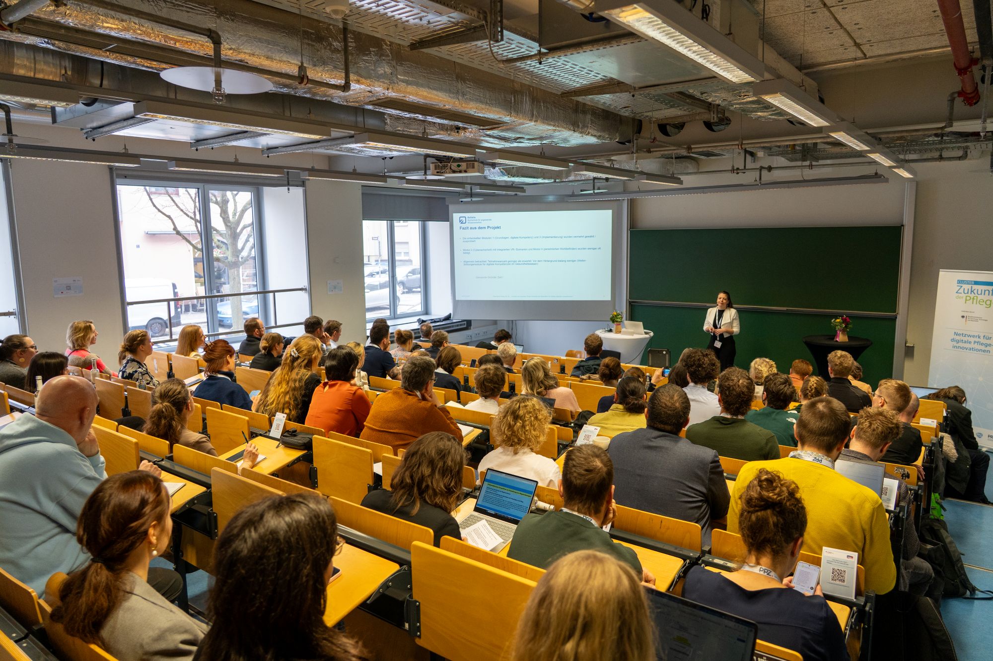 Vortragende steht vor einer Leinwand und spricht zu einem Publikum in einem Hörsaal mit Holzsitzreihen und großen Fenstern