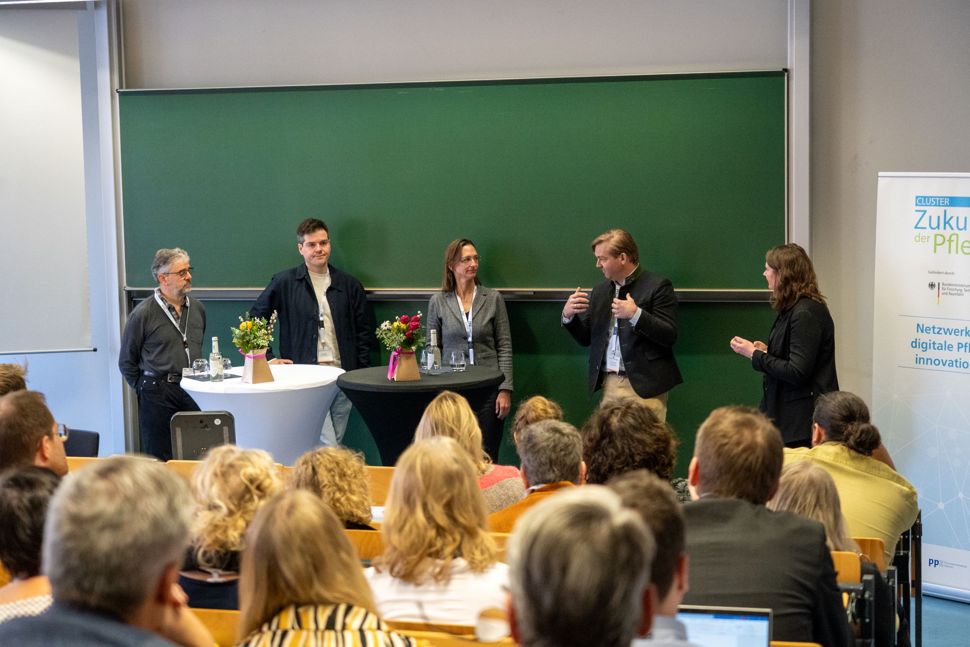 Fünf Personen stehen vor einer grünen Tafel und sprechen vor einem Publikum in einem Seminarraum