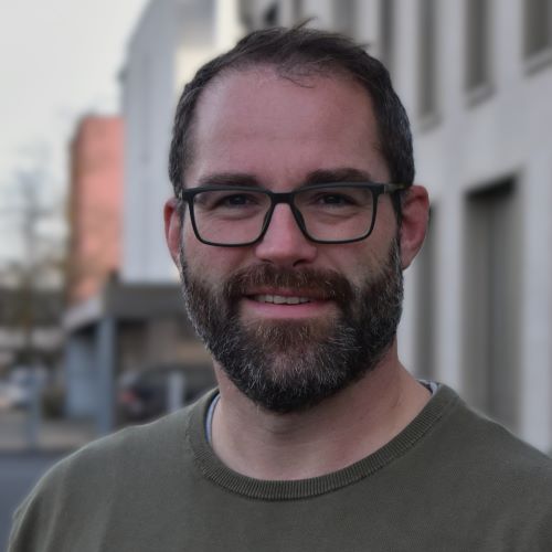 Das Bild zeigt einen Mann mit Brille und Bart. Er trägt einen olivgrünen Pullover und lächelt. Im Hintergrund sind Gebäude zu sehen, die leicht unscharf sind.