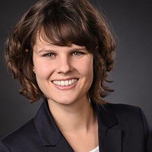 Professionelles Business-Porträt einer lächelnden Frau mit kurzen lockigen Haaren vor dunklem Hintergrund.