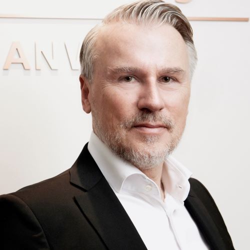 Mann mit grauem Haar trägt weißen Hemdkragen und schwarzen Anzug vor weißer Wand mit teilweise sichtbarem Schriftzug