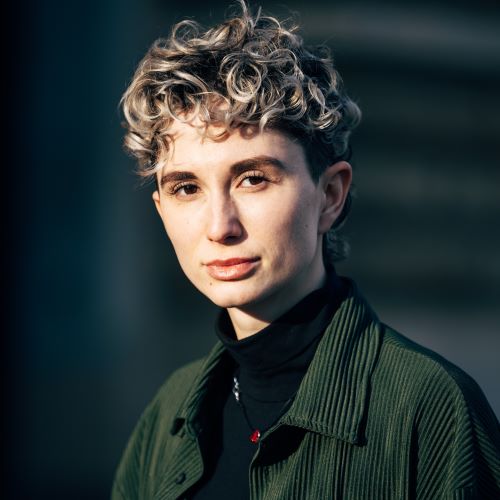 Person mit lockigem, hellen Haar trägt schwarzen Rollkragenpullover und dunkelgrüne Jacke vor unscharfem Hintergrund