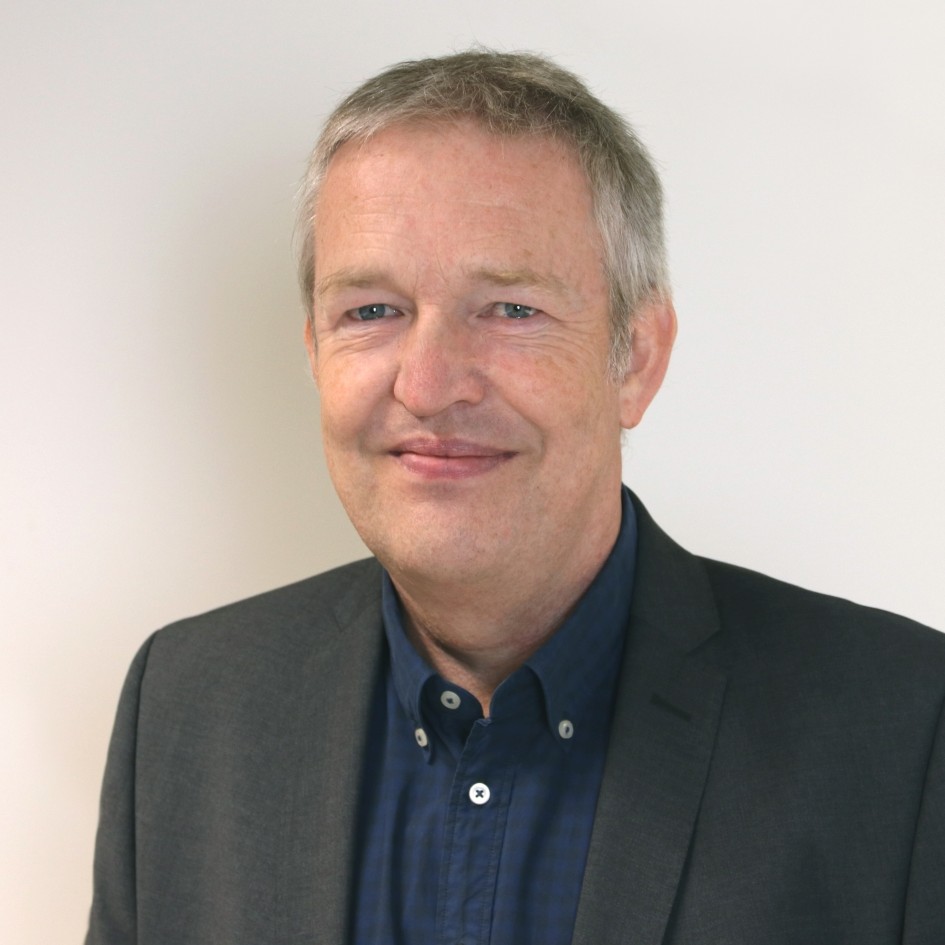 Person in dunklem Sakko und dunklem Hemd steht vor einer hellen Wand.