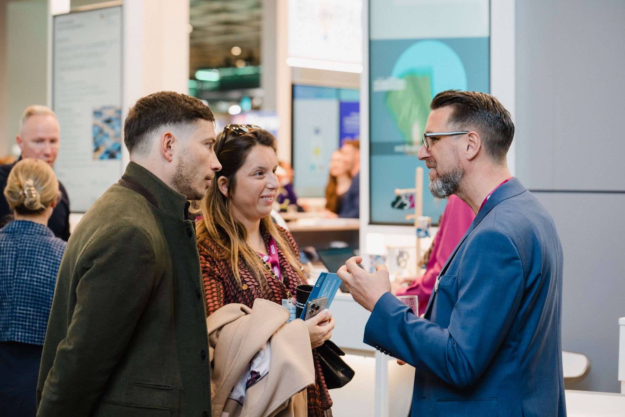 Drei Personen im Gespräch an einem Messestand, eine Person hält eine Tasse, zwei Personen tragen Mäntel und halten Broschüren