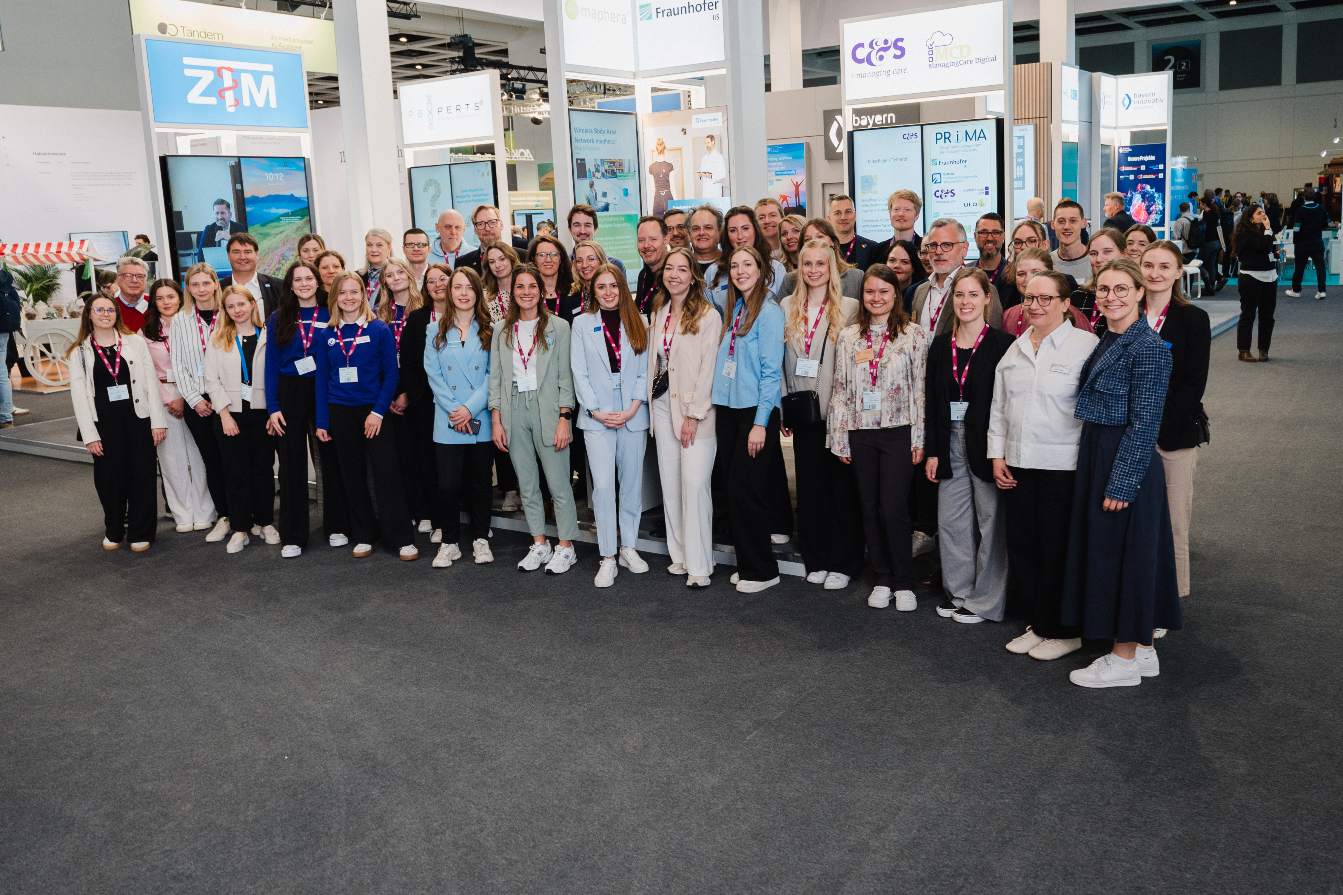 Große Gruppe von Menschen unterschiedlichen Alters und Geschlechts in einem Messe- oder Ausstellungsraum mit Ständen im Hintergrund