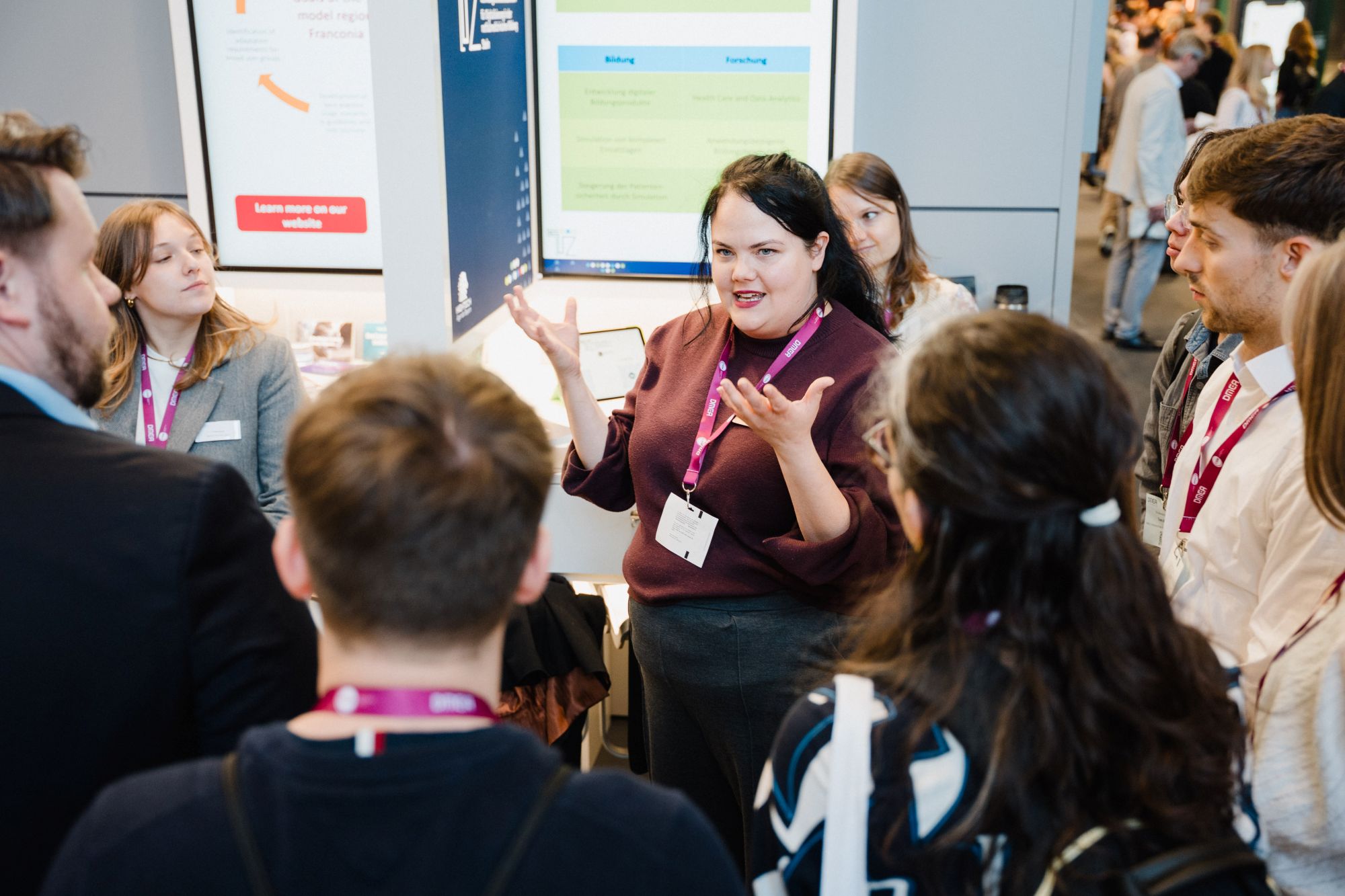 Frau mit lila Pullover und Namensschild spricht vor einer Gruppe von Menschen mit lila und roten Ausweisbändern in einem Messe- oder Konferenzraum