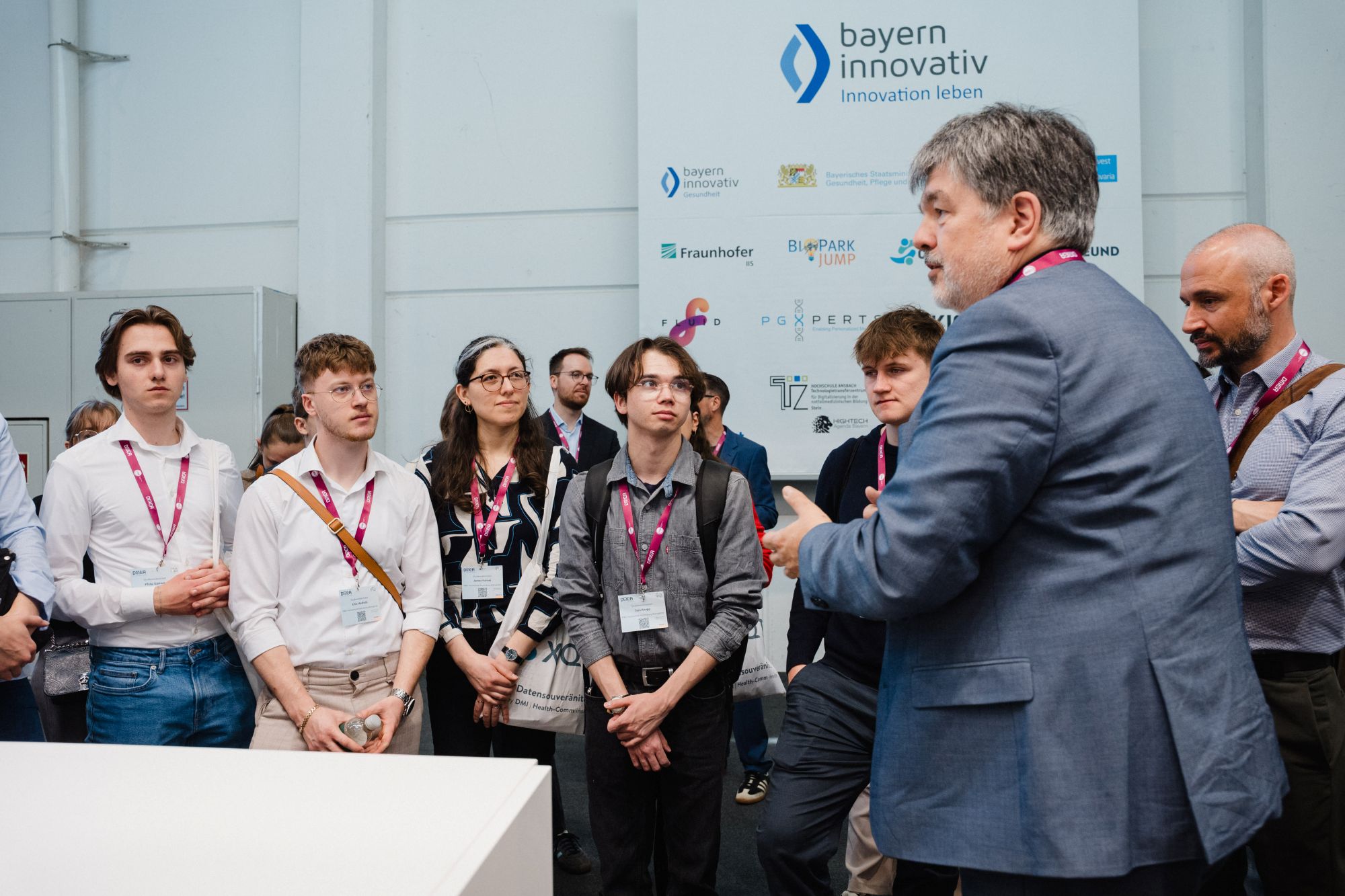 Gruppe von Menschen mit Namensschildern steht vor einem Mann im Anzug, der spricht, im Hintergrund Banner mit Logos von bayern innovativ und Partnern