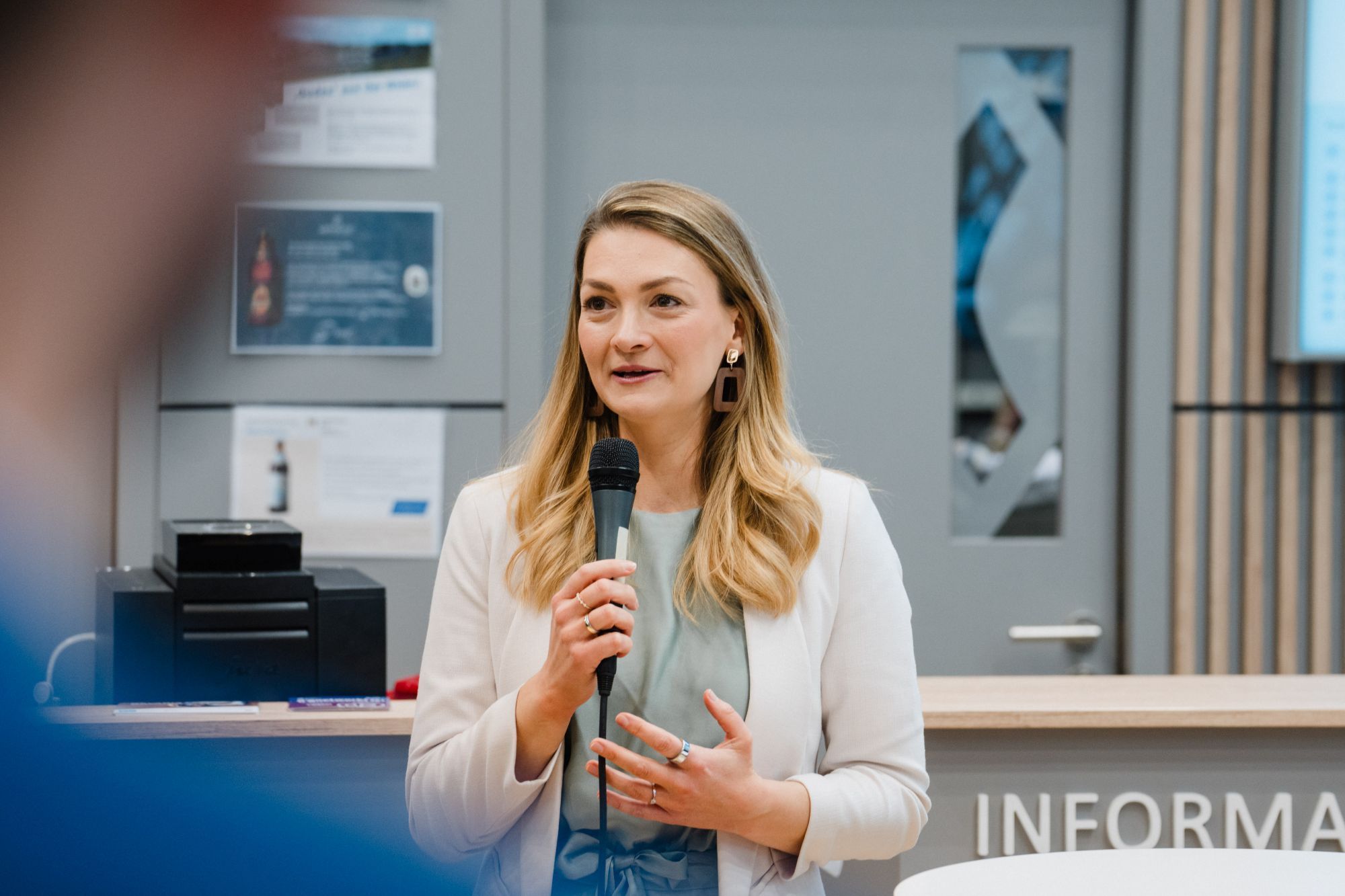 Frau mit langen blonden Haaren hält Mikrofon und spricht vor einem Informationsschalter.