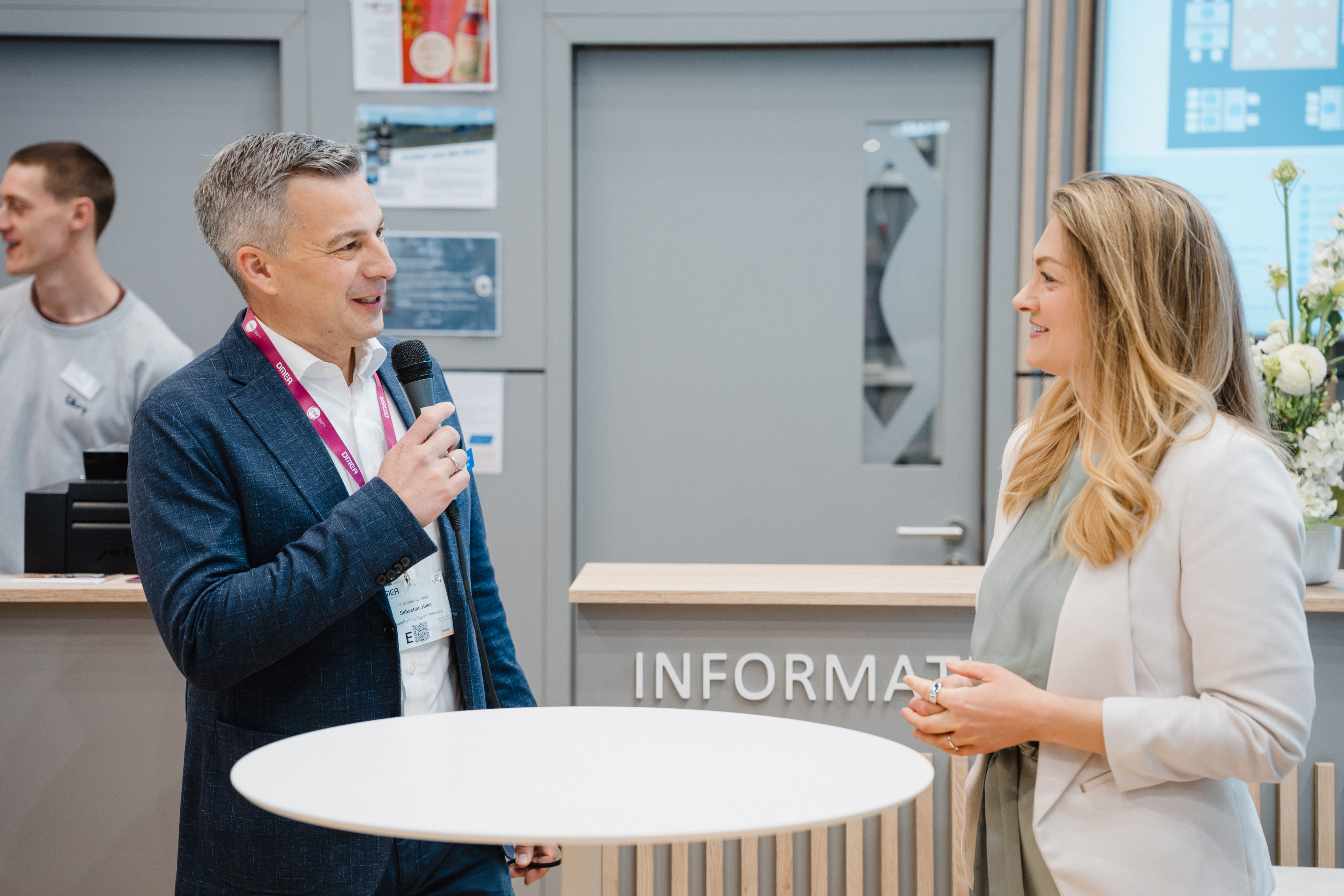 Mann mit Mikrofon im Gespräch mit Frau vor einem runden Stehtisch in einem hellen Raum mit Informationsschild im Hintergrund