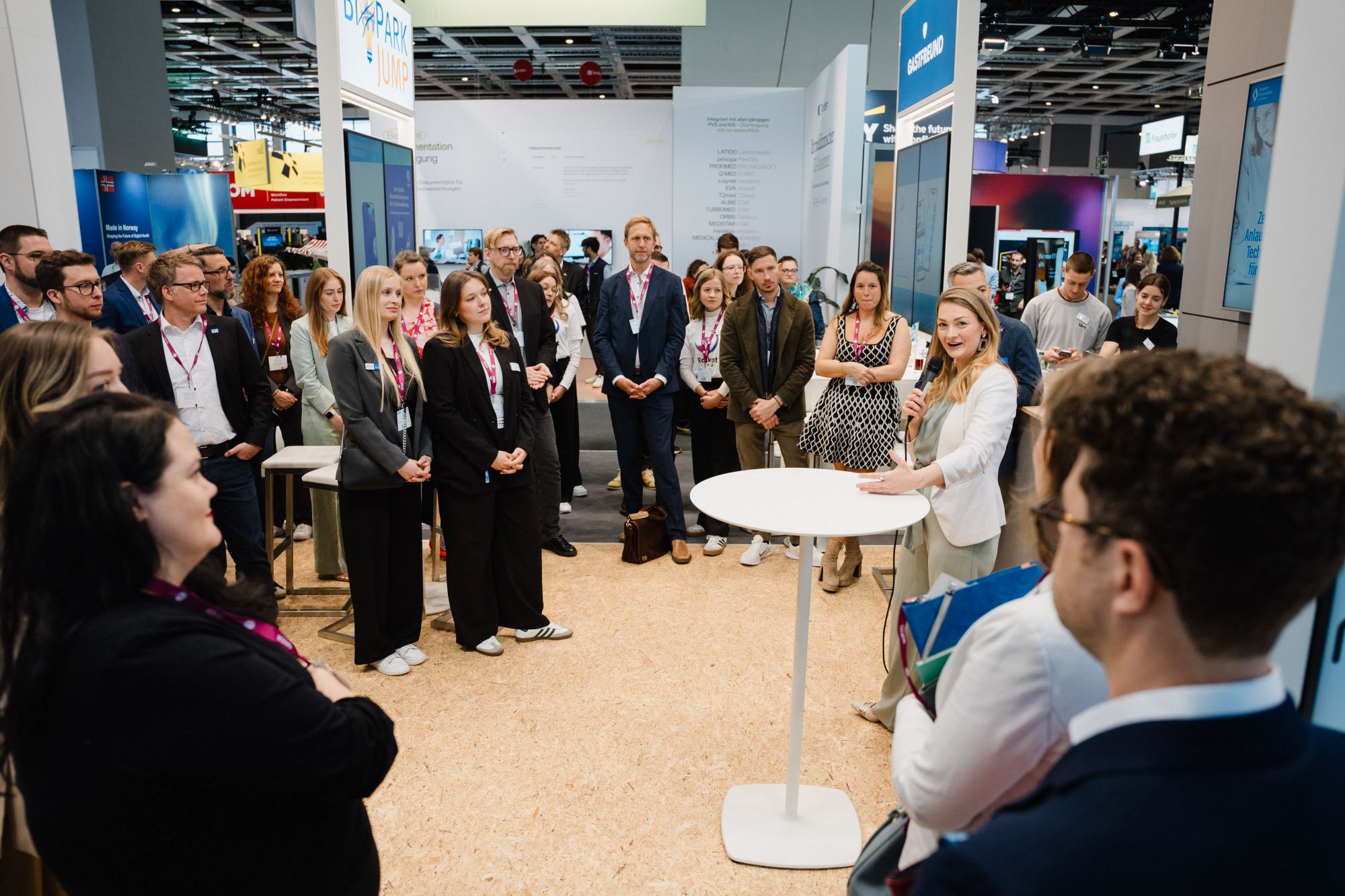 Frau spricht vor einer Gruppe von Menschen in einem hellen, modernen Messestand mit weißen Wänden und Stehtischen