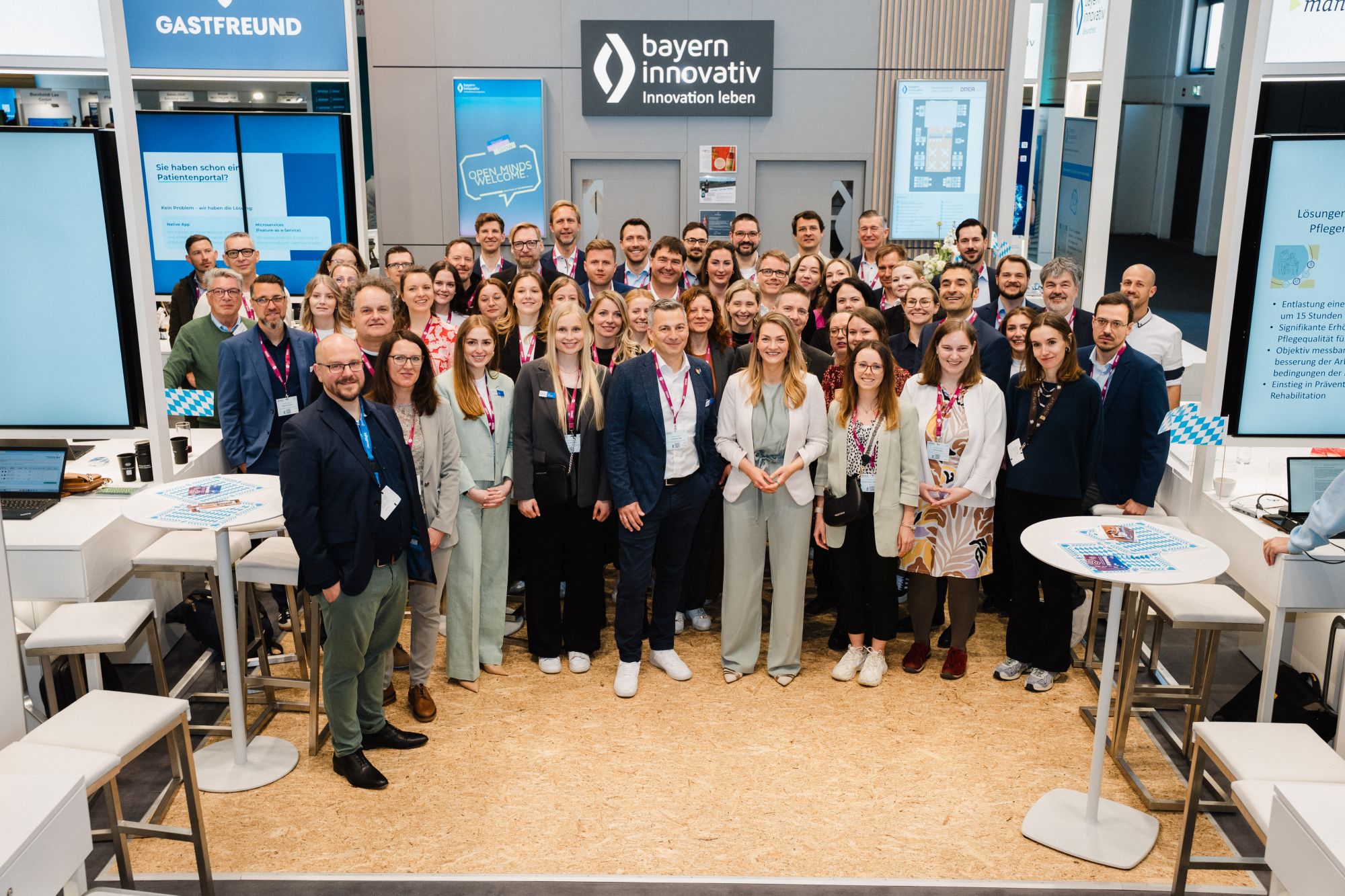 Große Gruppe von Menschen in einem modernen Messestand mit dem Logo von bayern innovativ an der Wand